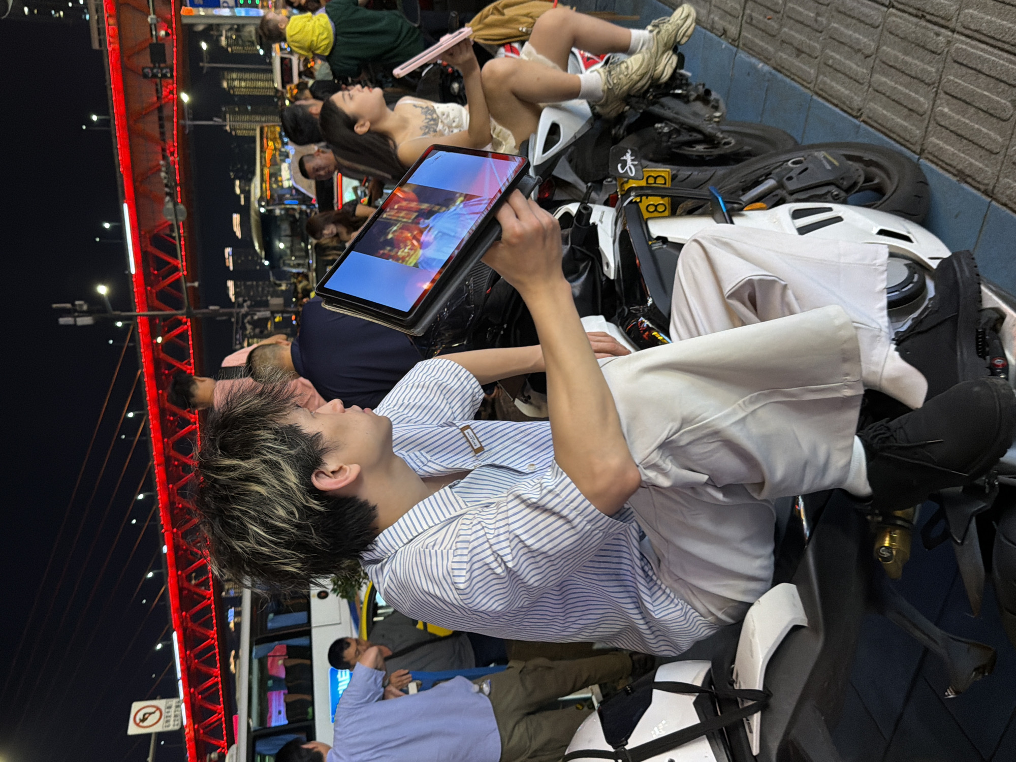 A young person with spiky, bleached hair sits on a motorcycle, holding a tablet displaying an image. They wear a blue and white striped shirt and light-colored pants. Next to them, a woman in a white dress sits on another motorcycle, looking away. In the background, people and vehicles are visible under a brightly lit red bridge. The scene is at night, with city lights in the distance.