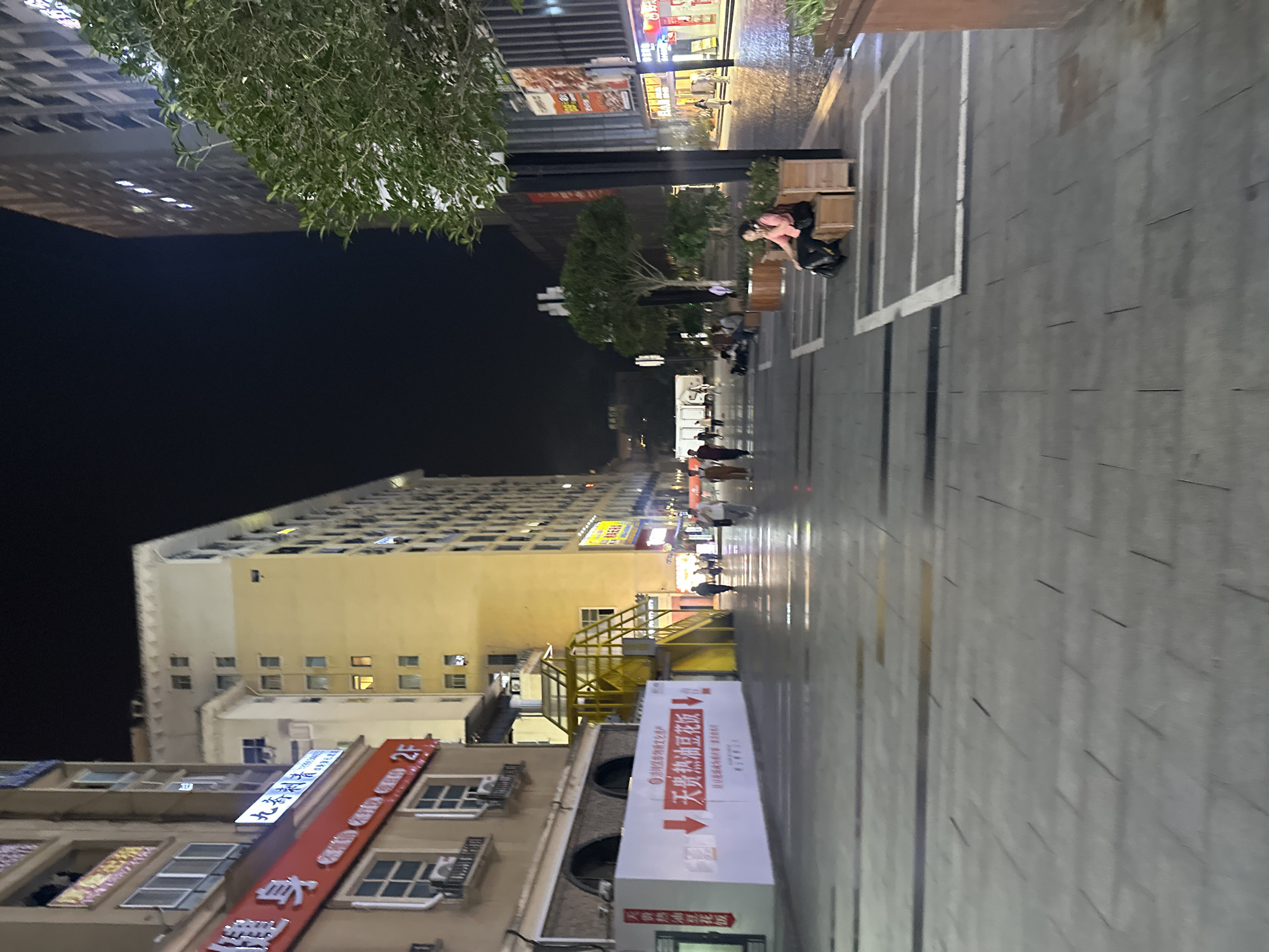 The image shows a street scene at night in the Chongqing High-tech Zone, China. The street is paved with stone tiles, and there are buildings on either side. A large white banner with red text is displayed on a building on the left. Several people are walking on the street, and some are sitting on benches. A tree is visible on the right side of the image. The overall impression is one of a busy urban environment at night.