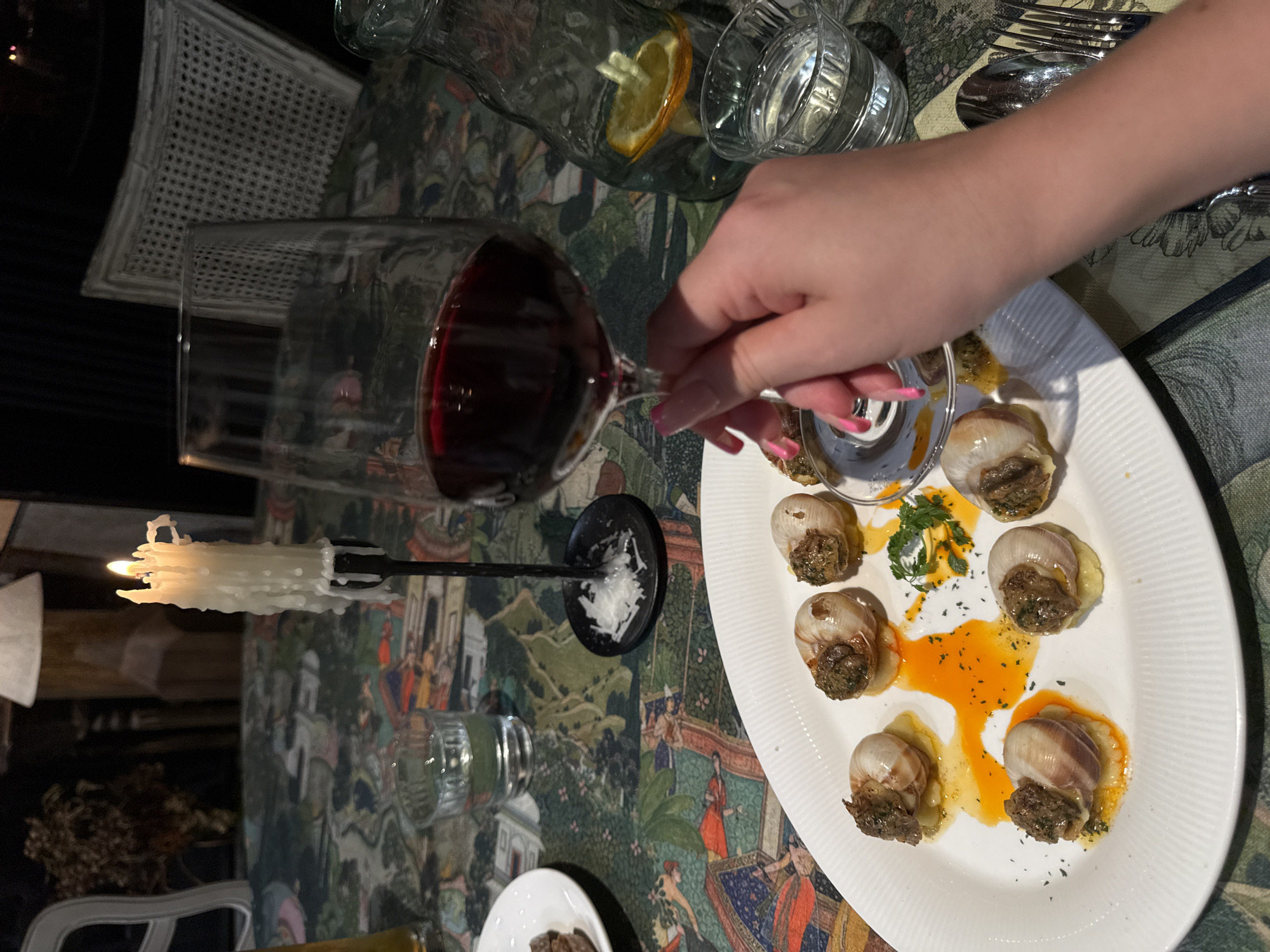 A high-angle, indoor shot depicts a dining experience. An oval, white plate displays escargots, garnished with herbs and bathed in an orange-yellow sauce. A hand with pink nail polish reaches for a snail fork resting within one of the shells. A glass of red wine lies horizontally on the table alongside a candle in a black candlestick, a water pitcher with citrus, and a glass of water. Silverware is placed at the top of the scene. The tablecloth has a patterned design. The location and city cannot be determined from the given image.