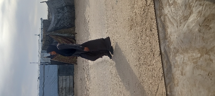 A person wearing dark clothing and a head covering walks across a sandy, uneven surface. In the background, there is a makeshift camp with various tents and coverings, including some adorned with decorative patterns. A low horizon line is visible under a cloudy sky. The overall scene conveys a sense of displacement or living in a temporary settlement. A wooden wall is on the right side of the image.