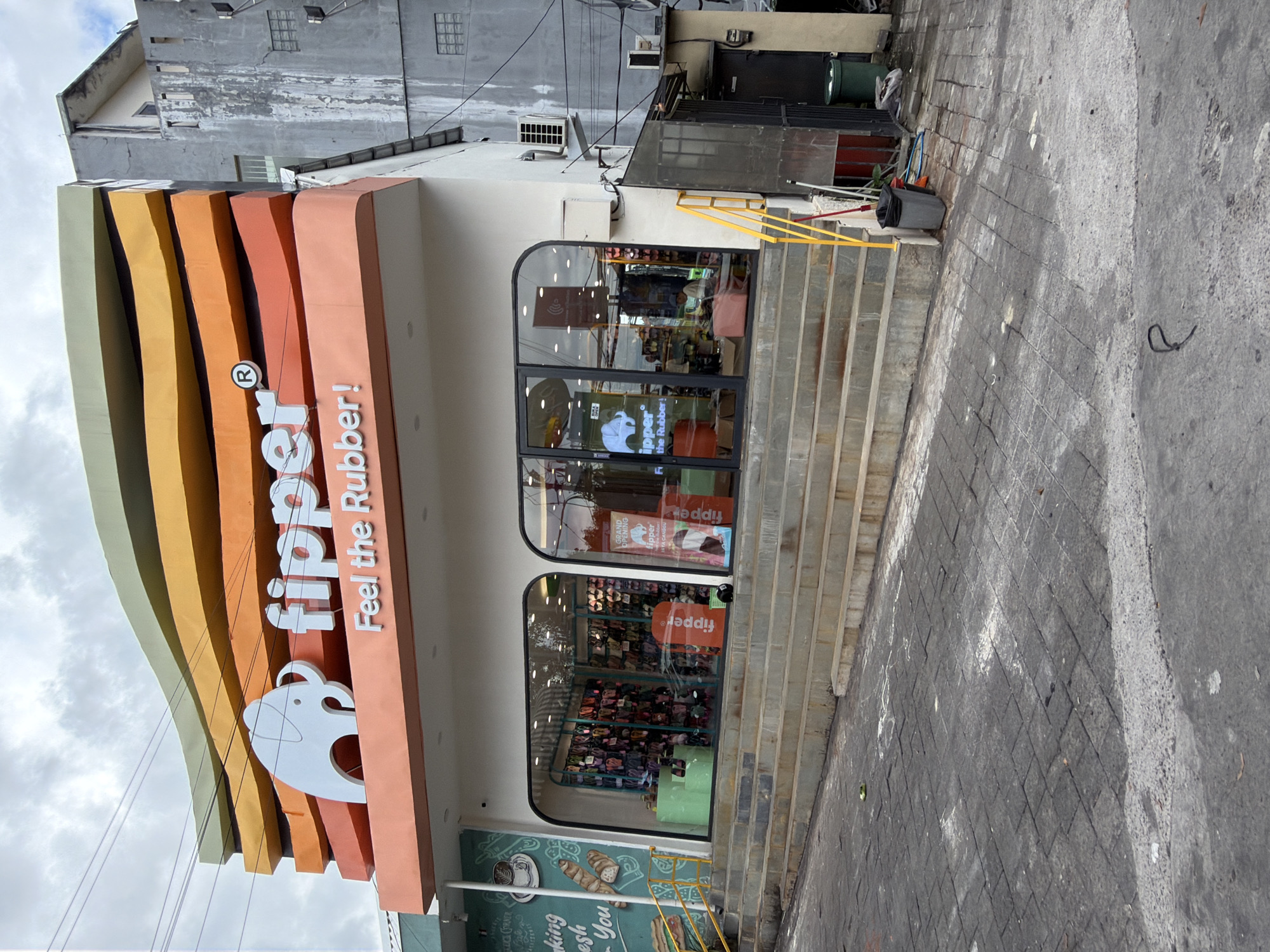 The image captures the exterior of a "fipper" store, a brand known for its rubber sandals, located in North Kuta, Indonesia. The store's facade is characterized by vertical, rounded segments painted in a gradient of colors, ranging from yellow-green to peach, with the "fipper" logo and the slogan "Feel the Rubber!" prominently displayed. A white, stylized hippo silhouette is part of the logo.
The store has large, arched windows showcasing the interior where displays of sandals can be seen. A set of stairs leads up to the store's entrance. To the right of the store is a brick paved sidewalk. A sign advertising "Kage Corner" is visible next to the store, featuring food items like coffee and pastries. The upper portion of the building behind the store appears to be an older, multi-story structure with visible signs of wear. Overhead, utility lines are visible against a cloudy sky. A gray trash can is located to the right of the store and near the steps.