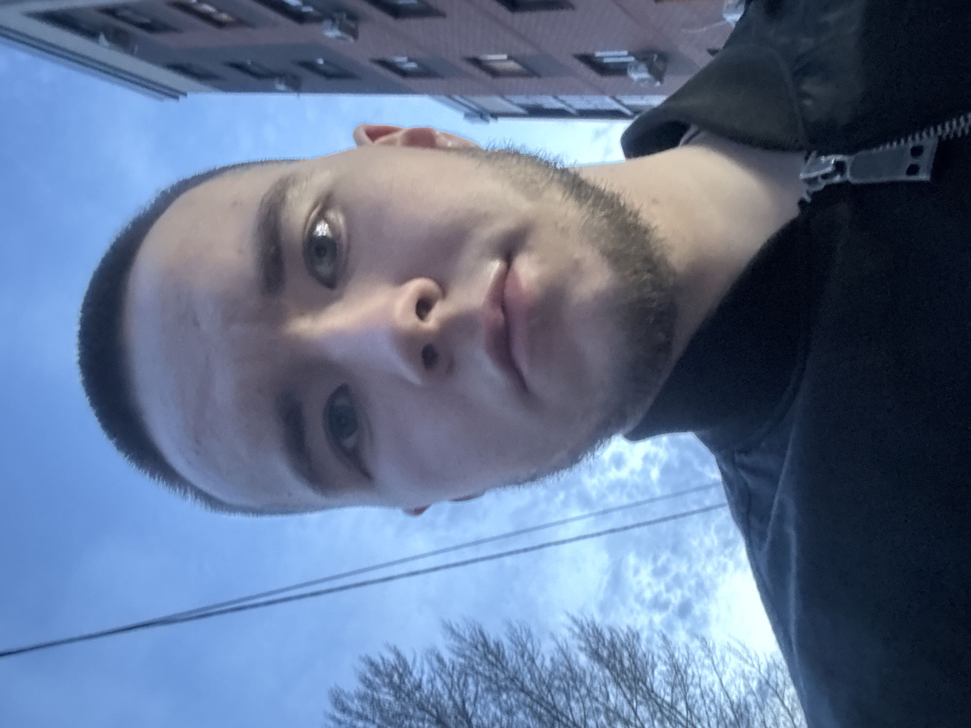 A selfie image shows a person with short, dark hair and a beard, wearing a dark-colored hooded jacket. The background features a building with multiple windows and a sky with clouds and bare tree branches. Electrical wires are visible against the sky. The location cannot be verified.