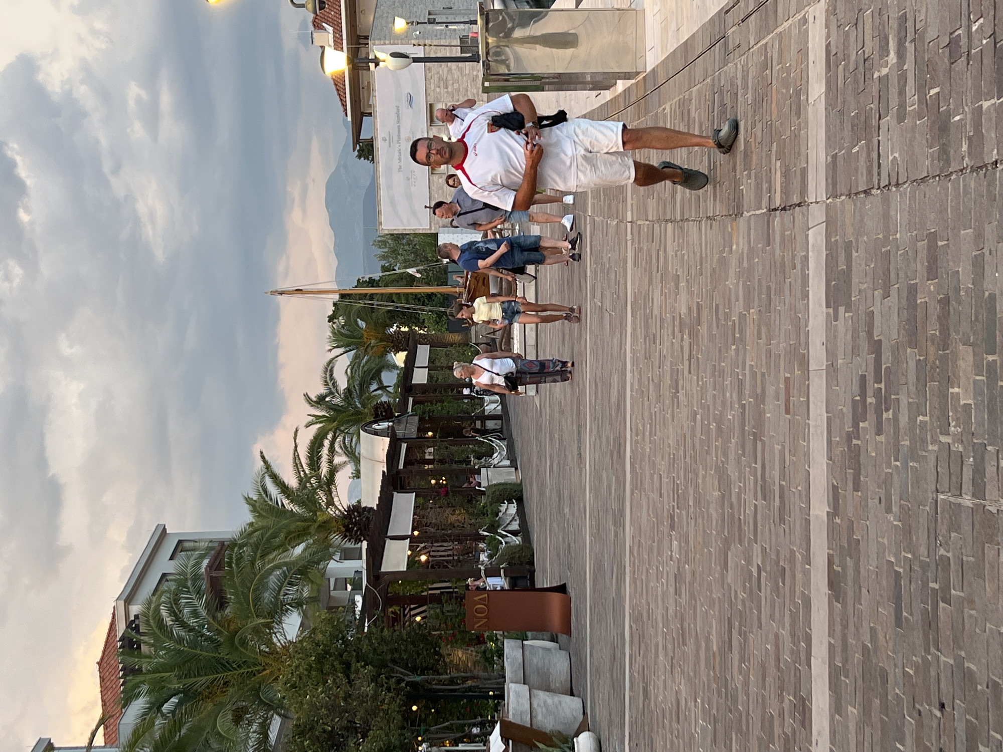 The image shows a scene on a paved walkway, likely a promenade, in Đenovići, Montenegro. The walkway is constructed with rows of brick-like paving stones. Several people are visible, appearing to be walking or standing.
In the foreground, a man is walking towards the camera, wearing glasses, a white polo shirt with red accents, and white shorts. He has a dark bag slung across his chest. Behind him, a few other individuals are walking in the same direction, including a man in a gray t-shirt and shorts, and another in a blue t-shirt and shorts. Further back, a woman in a yellow top and denim shorts is visible, as is another woman in a white top.
The setting includes buildings with balconies and awnings, some with white canopies. Palm trees are prominent in the scene, suggesting a warm climate. In the background, there are glimpses of hills or mountains under a cloudy sky, with the soft light indicating it is likely late afternoon or early evening. The overall mood appears relaxed and casual, consistent with a vacation or leisurely stroll in a coastal town.
There is visible text on a brown sign in the middle ground, appearing to read "VON" in white letters. The environment suggests a waterfront area, possibly near a harbor, with the presence of what looks like a mast from a sailboat.