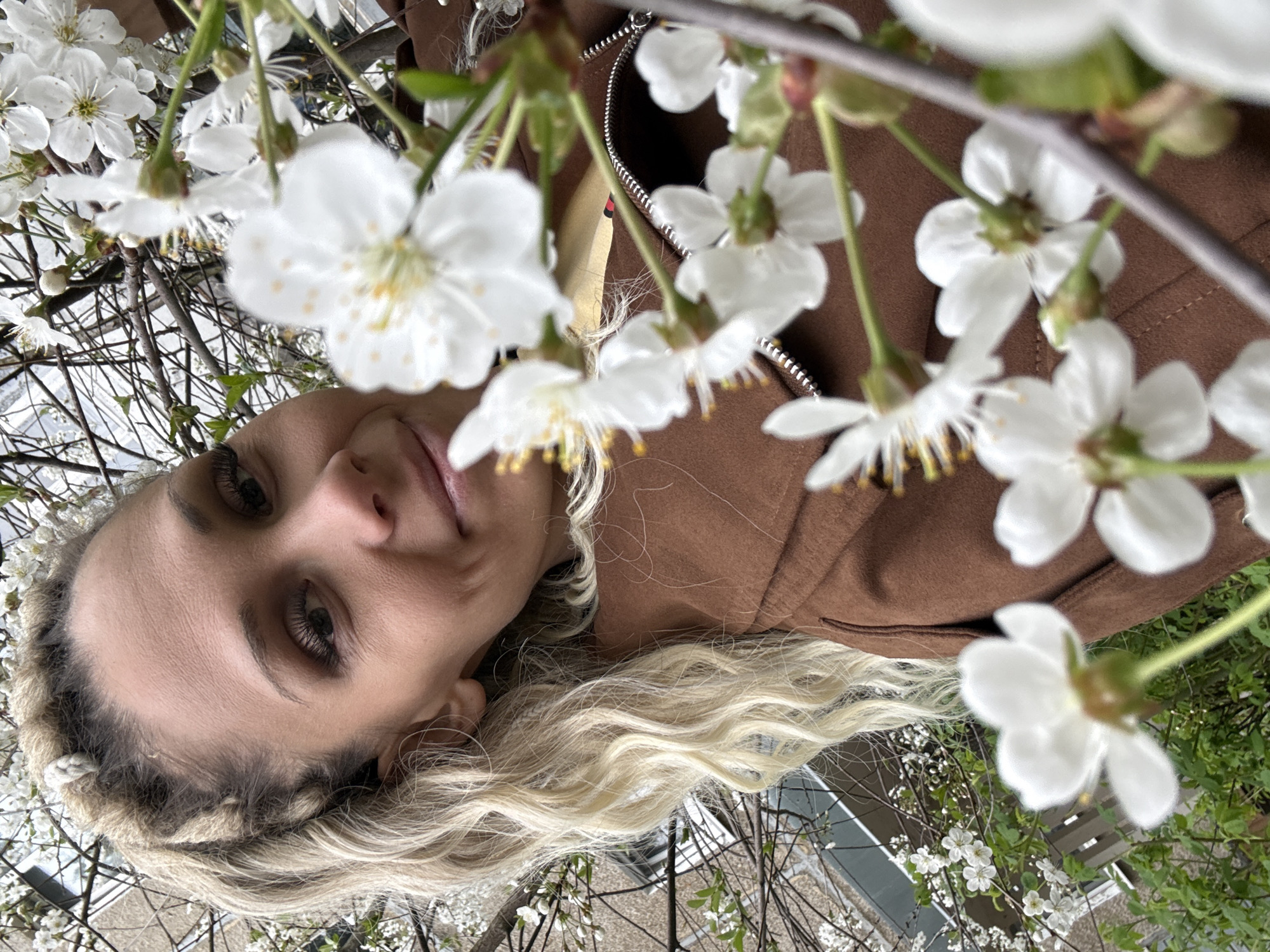 A woman with blonde, braided hair and a brown jacket is viewed from below, surrounded by flowering branches. The location appears to be outdoors, potentially in a garden or park within the city of Unknown, Unknown. The woman is looking down at the camera. The branches are covered in white blossoms.