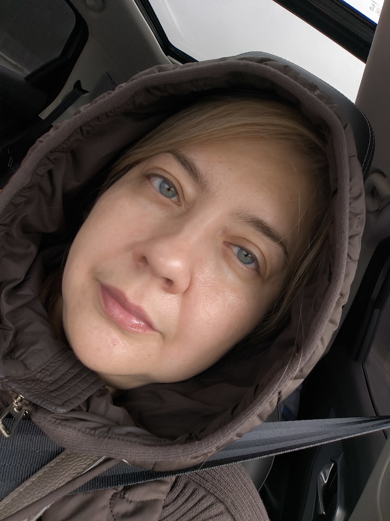 A close-up, angled portrait shows a woman taking a selfie inside a car. Her face is framed by the dark brown or grey hood of her padded jacket. She has blonde hair, light blue eyes, and her lips are painted with a light pink, glossy shade. Her expression is neutral to slightly contemplative.
The background reveals parts of a vehicle's interior, including a dark headrest behind her, a light-colored sunroof above, and a dark seatbelt visible across her chest and shoulder. The lighting is soft and diffused, suggesting it is daytime. The hooded jacket implies cooler weather. There is no visible text in the image.