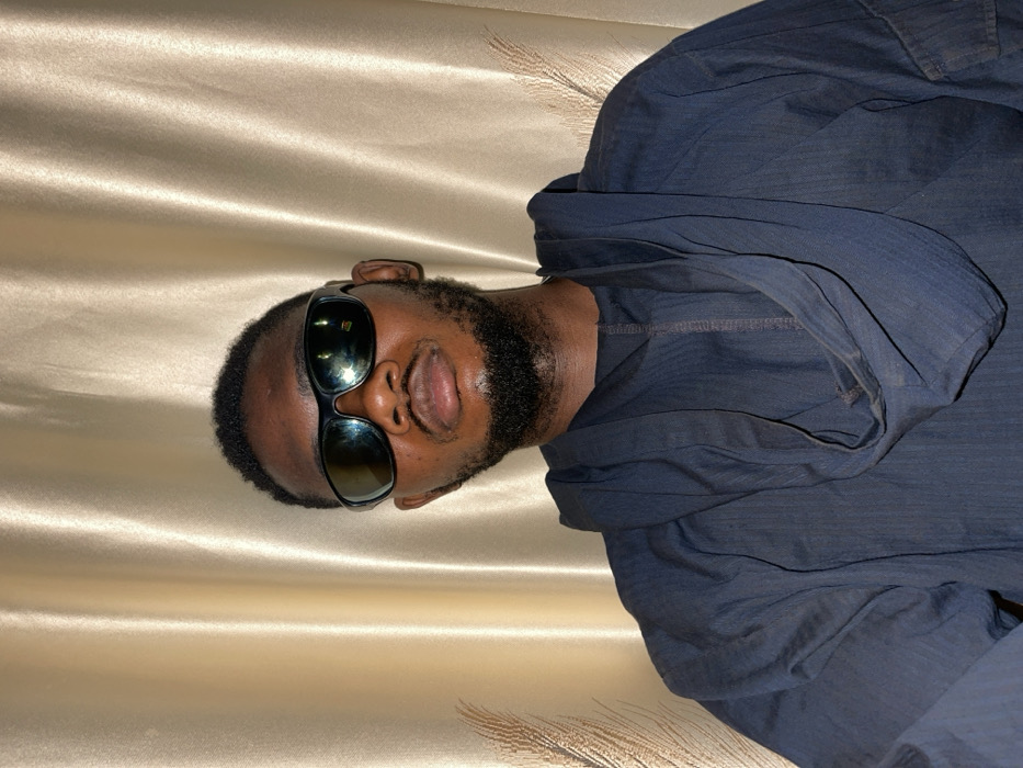 A man with dark skin, a black beard, and sunglasses faces forward. He wears a dark blue, hooded shirt. The background is a shiny, golden fabric draped vertically. The man's head is tilted to his right. The image is rotated to the left by 90 degrees.