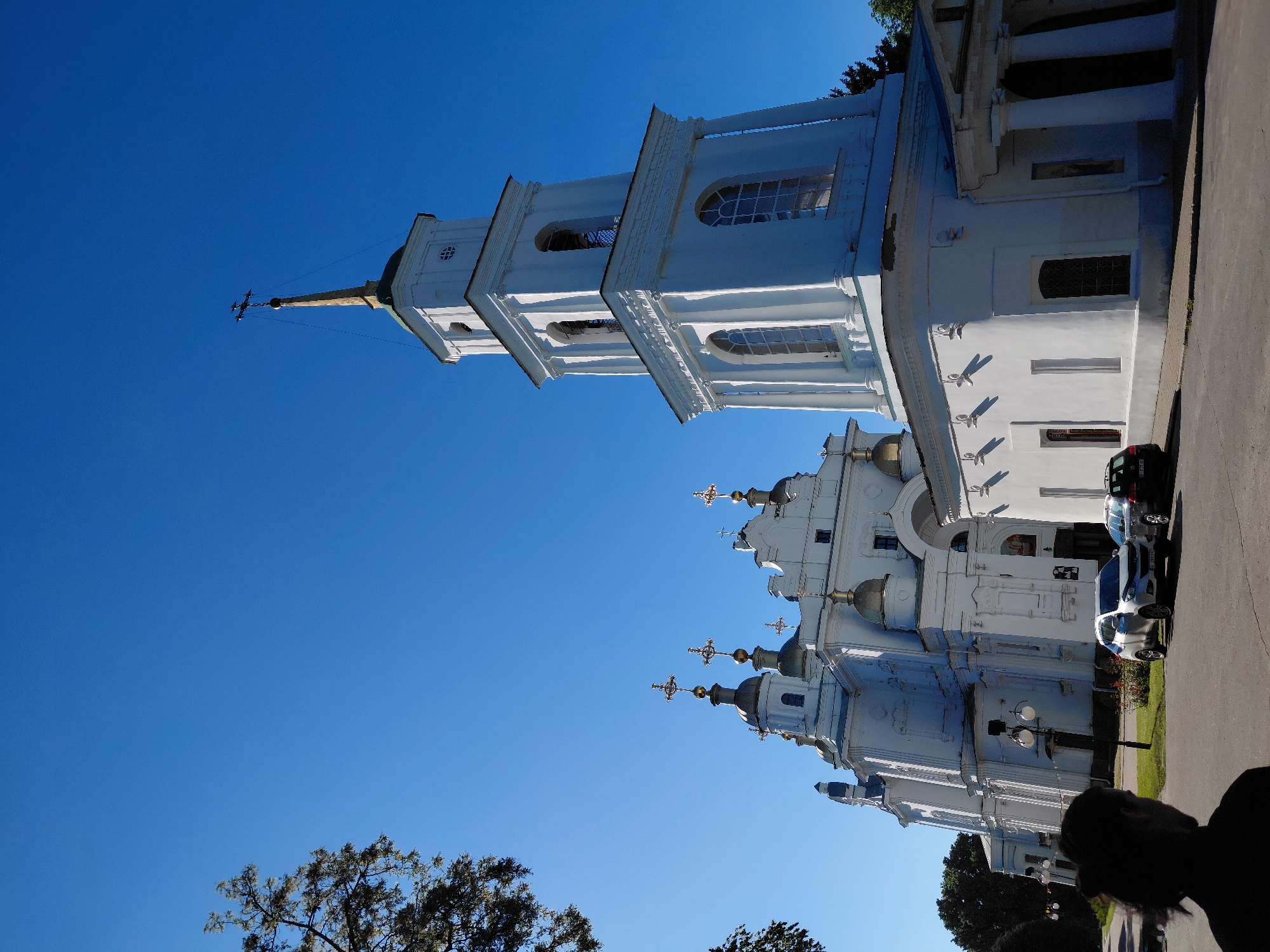 The image captures a large, ornate white church or cathedral under a clear, bright blue sky in Poltava, Ukraine. The main subject is the impressive ecclesiastical building, which features multiple tiers, classical architectural elements like columns and cornices, and numerous arched windows.
A tall, multi-tiered bell tower or spire, also white, dominates the left side of the frame, culminating in a golden spire topped with a cross. To the right, the main body of the church is visible, adorned with several green-domed cupolas, each crowned with a distinctive golden Orthodox cross. The architecture suggests a Baroque or Neoclassical style.
The scene is bathed in bright sunlight, indicated by sharp shadows cast by the church's architectural details on its white walls and by elements like a lamppost on the right. The time of day appears to be either morning or afternoon due to the strong, direct sunlight. The sky is completely clear, indicating fair weather.
On the right side of the church, a paved area or road is visible, along which two cars are parked. One is a dark-colored SUV or van, and the other is a lighter-colored hatchback. A partial silhouette of a person's head and shoulders is seen in the lower right corner, suggesting someone is standing nearby. Some trees and green vegetation are visible in the background and foreground, particularly on the lower left and upper right corners.
Visible text includes partial license plates on the parked cars: "AA 3072 CK" on the lighter car, and "AE..." on the darker car. No other legible text is apparent on the church building itself.
