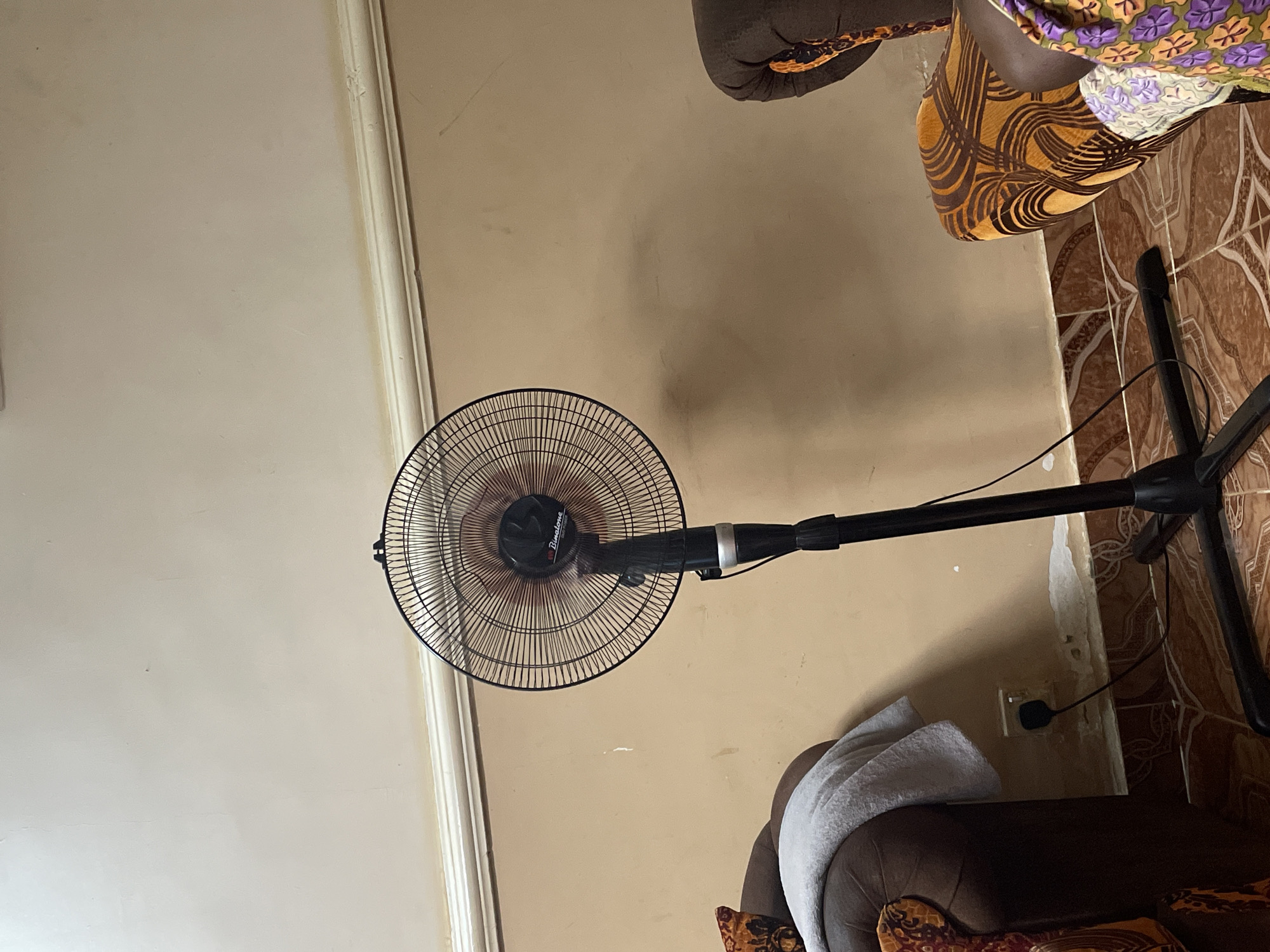 This image captures an indoor scene in Narayi, Nigeria, depicting a living space.
A black Binatone pedestal fan is centrally positioned, operating with visible blade blur. The fan features a black circular grille and stands on a stable black cross-shaped base.
To the left, a dark brown upholstered sofa or armchair is partially visible, draped with a light grey towel. Beneath the sofa, a white wall outlet is visible on the light brown wall, with a black plug and cord connected, presumably powering the fan. The wall exhibits signs of wear, including peeling paint or plaster near the floor, revealing a lighter subsurface.
To the right, a portion of another upholstered furniture item is visible, featuring a distinct orange and dark brown swirling geometric pattern. A person's lower body, clothed in a vibrant purple, orange, and green floral-patterned fabric, is partially visible at the extreme right edge, adjacent to the patterned furniture.
The floor throughout the scene is covered with brown and tan patterned ceramic tiles. The light brown walls are topped with a white crown molding, separating them from the off-white ceiling. Minor scuff marks and discolorations are present on the walls and ceiling.