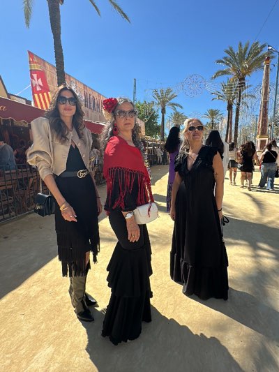 The image captures three women posing outdoors in Jerez de la Frontera, Spain, during a local celebration. The woman on the left wears a short black dress with fringe, a beige bomber jacket, black boots, and sunglasses. The woman in the middle is dressed in a long black dress with tiered ruffles, accessorized with a red shawl, a red flower in her hair, and sunglasses. The woman on the right wears a long black dress with ruffles. They are standing in a light-colored outdoor area, possibly a plaza or fairground, with palm trees and decorated structures in the background. People can be seen walking in the background. The sky is clear and blue.
