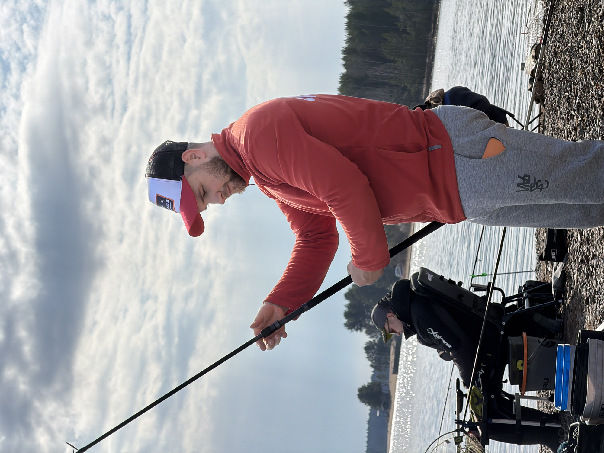 This photograph captures two men engaged in fishing by a body of water in Chaykovsky, Russia, under a partly cloudy sky. The scene is set on a rocky or gravelly shoreline, with a dense line of trees visible on the opposite bank.
In the foreground, a man with a light beard, wearing a red hooded long-sleeved top, grey sweatpants, and a black, white, and red baseball cap, is leaning forward, focused on the water. He holds a fishing rod with both hands, appearing to be casting or adjusting his line. The white panel of his cap has a logo and text, possibly "ENGINS". The grey sweatpants feature stylized, indistinct characters. The fishing rod he holds also appears to have text, possibly "MAVERIX".
Further back and to the right, a second man is seated on a specialized fishing chair or platform, also facing the water. He wears a dark jacket with "MAVER" visible on the sleeve, dark pants, and a dark cap. He is surrounded by an extensive fishing setup, including multiple fishing rods, a net, and various buckets. One blue bucket is labeled "BARU", and another piece of equipment clearly states "FEEDER", indicating the type of fishing being done.
The water surface is rippled, reflecting the bright sky. The overall atmosphere suggests a calm, focused outdoor activity during daytime, with comfortable weather conditions.