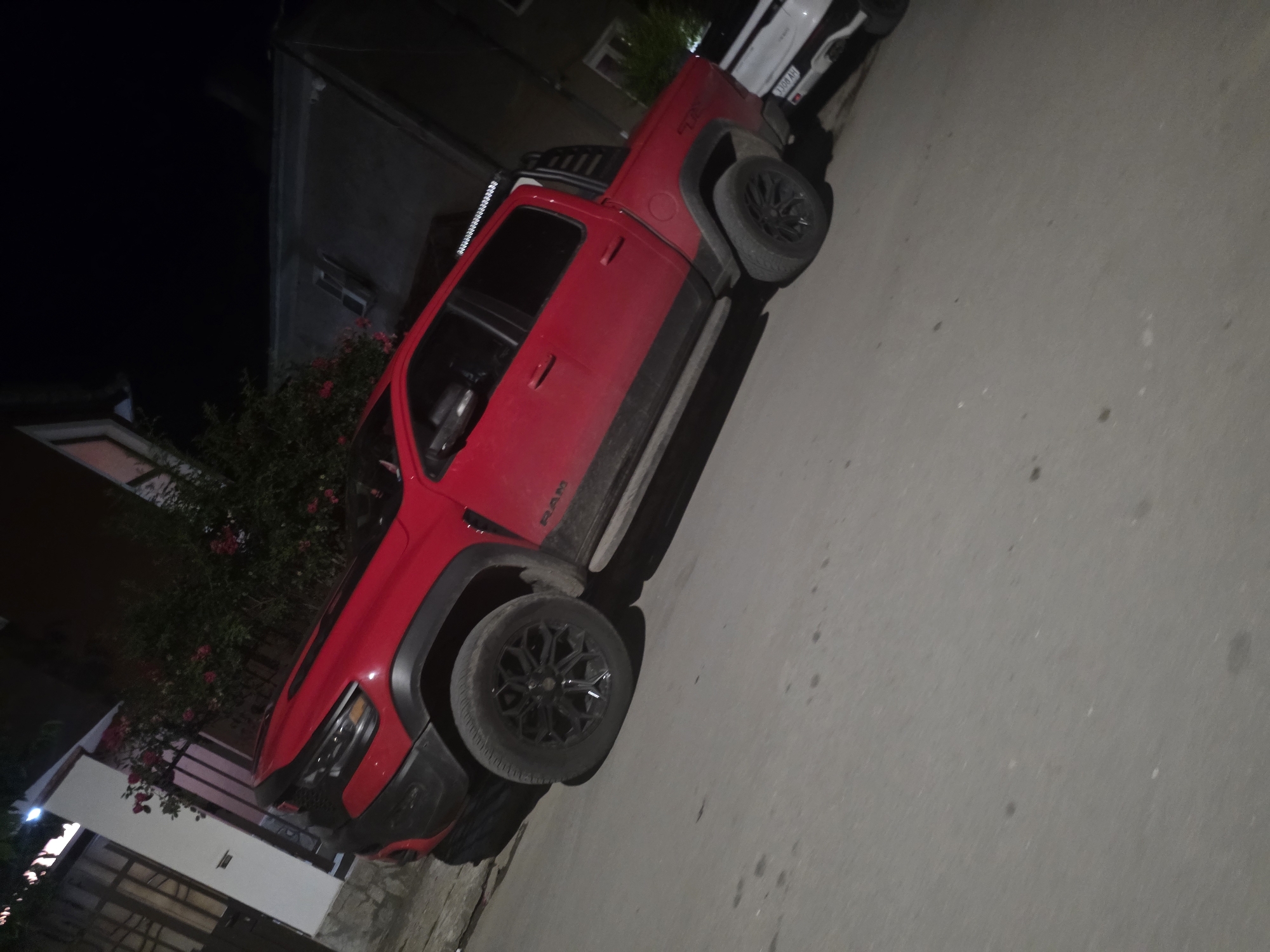 A red Ram TRX pickup truck is parked on a paved street at night in Targovishte, Bulgaria. The truck is angled, facing towards the left of the frame, with its front tires turned. The street is grey and appears to be illuminated by ambient light from nearby buildings and possibly streetlights. To the left of the truck, there is a white building with a fence and some greenery with red flowers. The truck has black wheels and black fender flares. The side of the truck displays the "RAM" logo. Further down the street, to the right of the truck, a white SUV is partially visible, also parked. The background consists of buildings that are dimly lit. The overall scene suggests a residential or semi-urban area at night.