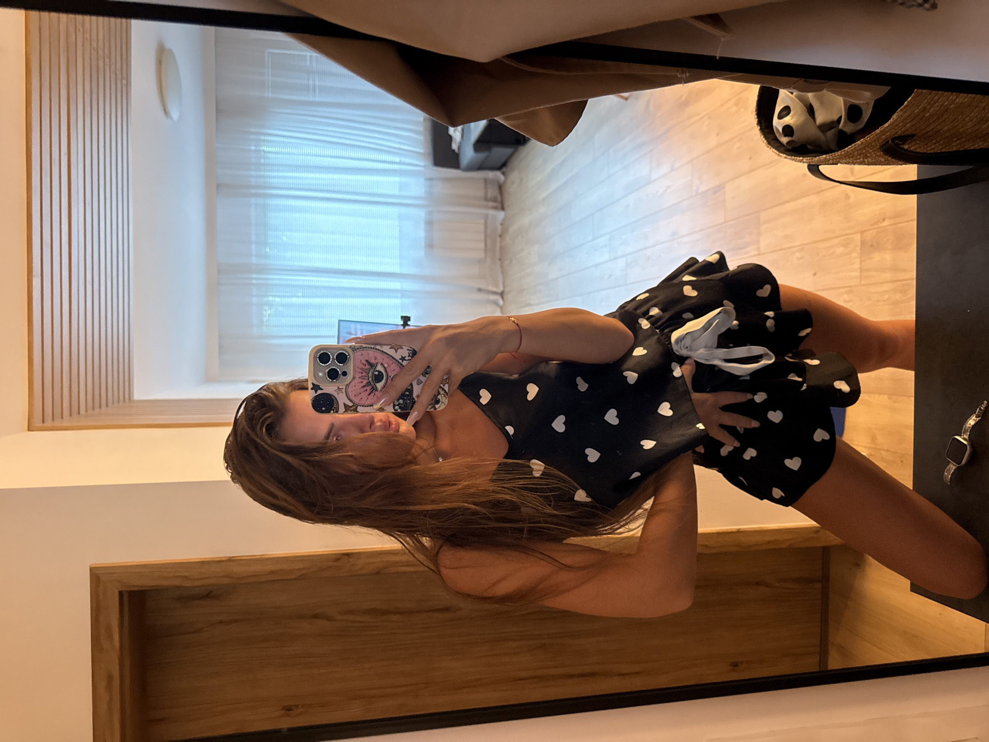 The media file shows a young woman taking a mirror selfie in what appears to be a bedroom or dressing area. The woman is wearing a black dress with small white hearts. Her hair is long and brown. She is holding a smartphone with a colorful case to take the photo.
In the background, reflected in the mirror, is a room with light-colored wooden floors and walls. There are vertical wooden slats on one wall, and a window with sheer blinds on another. A straw bag with a black trim and a white scarf with black polka dots is visible on a surface. A watch is also visible on a dark surface to the right.
The lighting suggests it could be daytime, possibly late afternoon or early evening, given the warm tones in the reflections. The overall mood appears casual and relaxed, as the woman is posing for a photograph in what seems to be a private space. There is no visible text in the image. The location context of Plovdiv, Bulgaria is provided but not visually evident in the image itself.