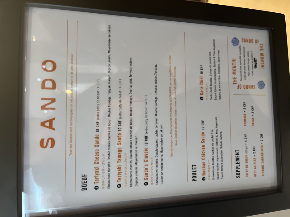 The image captures a menu board, likely illuminated, displaying food options. The setting appears to be indoors, possibly a restaurant or cafe, given the clear presentation of dishes and prices. No people are visible.
The menu is predominantly in French, with some Japanese katakana characters accompanying the dish names. The currency used is Swiss Francs (CHF), which aligns with the provided context of Genève, Switzerland.
The menu is organized into sections: "BOEUF" (Beef), "POULET" (Chicken), "SUPPLÉMENT" (Supplement), and a special "SANDO OF THE MONTH!".
Under "BOEUF", options include "Teriyaki Cheese Sando" (18 CHF), "Teriyaki Tamago Sando" (19 CHF), and "Sando's Classic" (18 CHF). These items feature toasted shoku-buns, double beef patties, various toppings such as cheese, teriyaki sauce, umami onions, takuan mayonnaise, fried egg, tomatoes, and green lettuce. An extra beef patty costs 4 CHF. A note indicates that any beef burger can be prepared vegetarian for a 2 CHF supplement, utilizing Swiss soy and plant-based patties.
The "POULET" section lists "Nanban Chicken Sando" (19 CHF) and "Kara-Chiki" (19 CHF). These sandwiches also use toasted shoku-buns and include fried chicken thigh, accompanied by different sauces (Nanban sweet-sour, Sichimi & Yuzu togarashi, Japanese tartare, gochujang, spicy mayo) and ingredients like sweet salad, cucumber pickles, and coleslaw.
The "SUPPLÉMENT" category offers additional items: an 80g beef patty (4 CHF), a fried egg (2 CHF), caramelized onions (2 CHF), cheese (2 CHF), and sauce (2 CHF).
The "SANDO OF THE MONTH!" section promotes a monthly special created by the team, stating that 1 CHF from each sale of this burger goes to the creator. The overall presentation is clean and functional, designed to inform customers about the offerings.