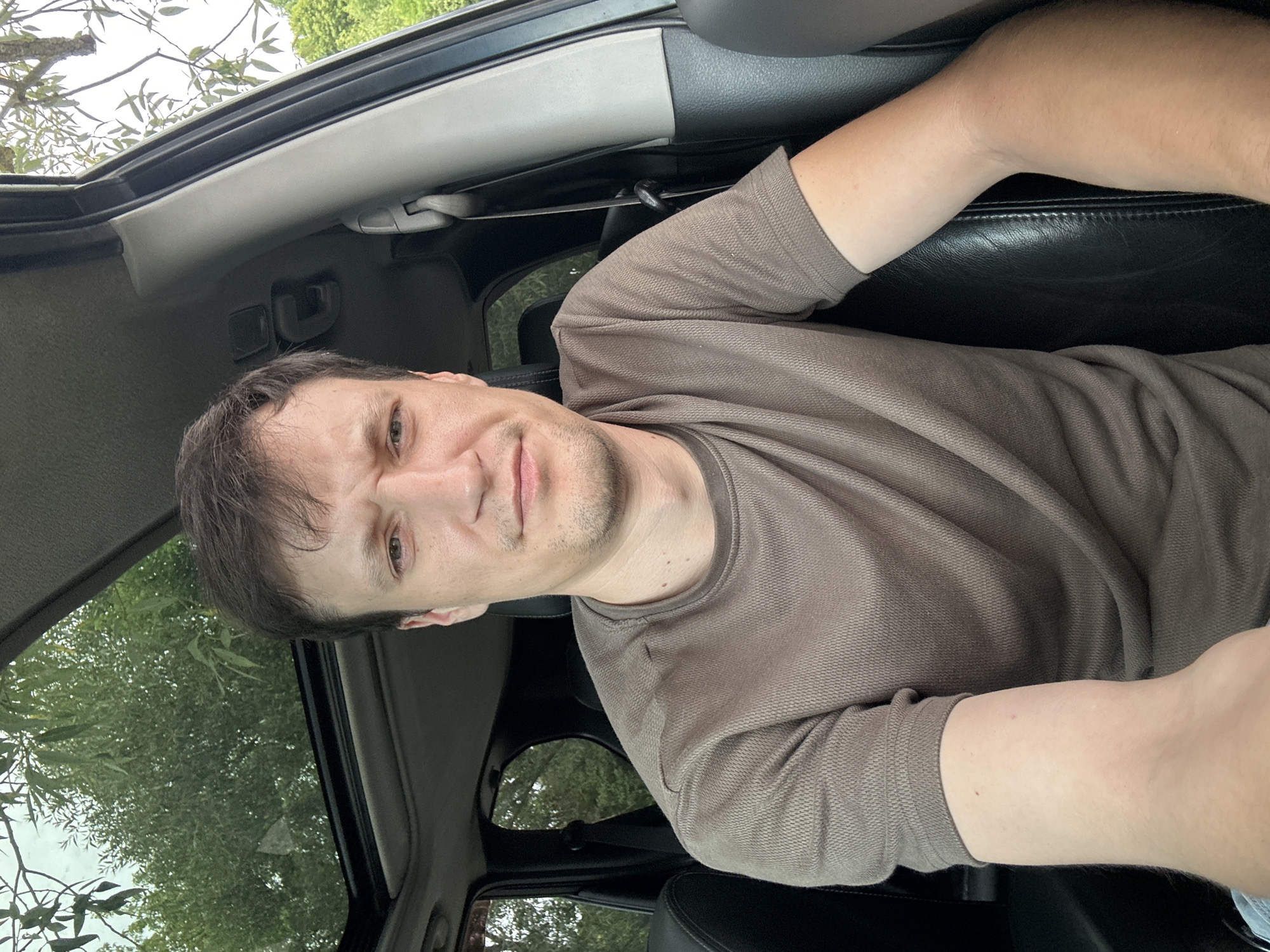 A man is sitting in the driver's seat of a car, looking at the camera. He has short brown hair, light skin, and is wearing a brown t-shirt. The car has a sunroof open, and trees and foliage can be seen through it and through the car windows. The interior of the car is visible, including black leather seats and a seatbelt. The scene is outdoors, likely during the daytime.