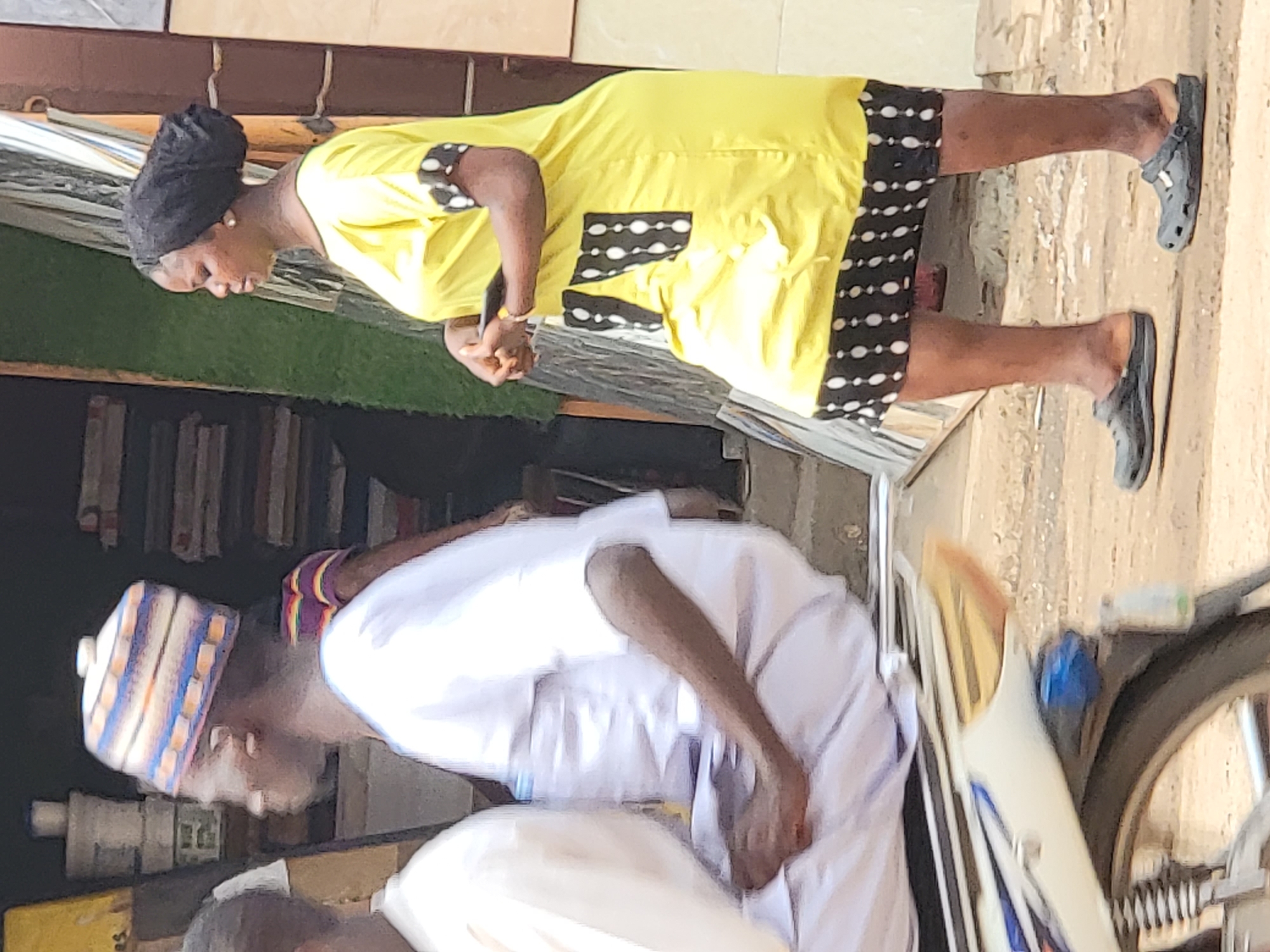 In Maiburiji, Nigeria, a woman wearing a yellow dress with black and white trim stands on a concrete surface near what appears to be a storefront with stacked items inside. She is looking down at her hands. She wears black flats. Nearby, a man in a white outfit and a striped cap is bent over near a bicycle or motorcycle.