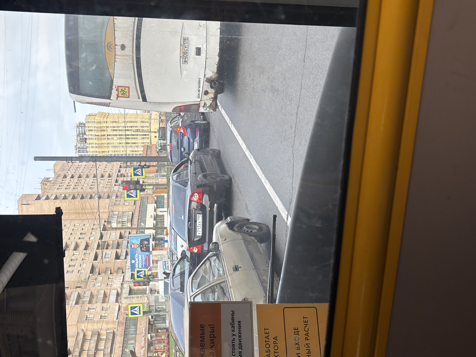 The image shows a view from inside a vehicle, possibly a bus or a tram, looking out at a street scene. A line of cars is visible in the adjacent lane, along with a large white bus, its rear bearing the license plate "B408CY 178". The license plates of other cars are visible, including "Y713XX 198". The buildings across the street are tall and densely packed, with a prominent banner promoting pride in Russia. There are pedestrian crossing signs with flashing red lights. The lower portion of the image shows a sign in Russian, partially obscured by the window frame and with text that includes a warning to passengers. The environment indicates an urban setting with traffic congestion.