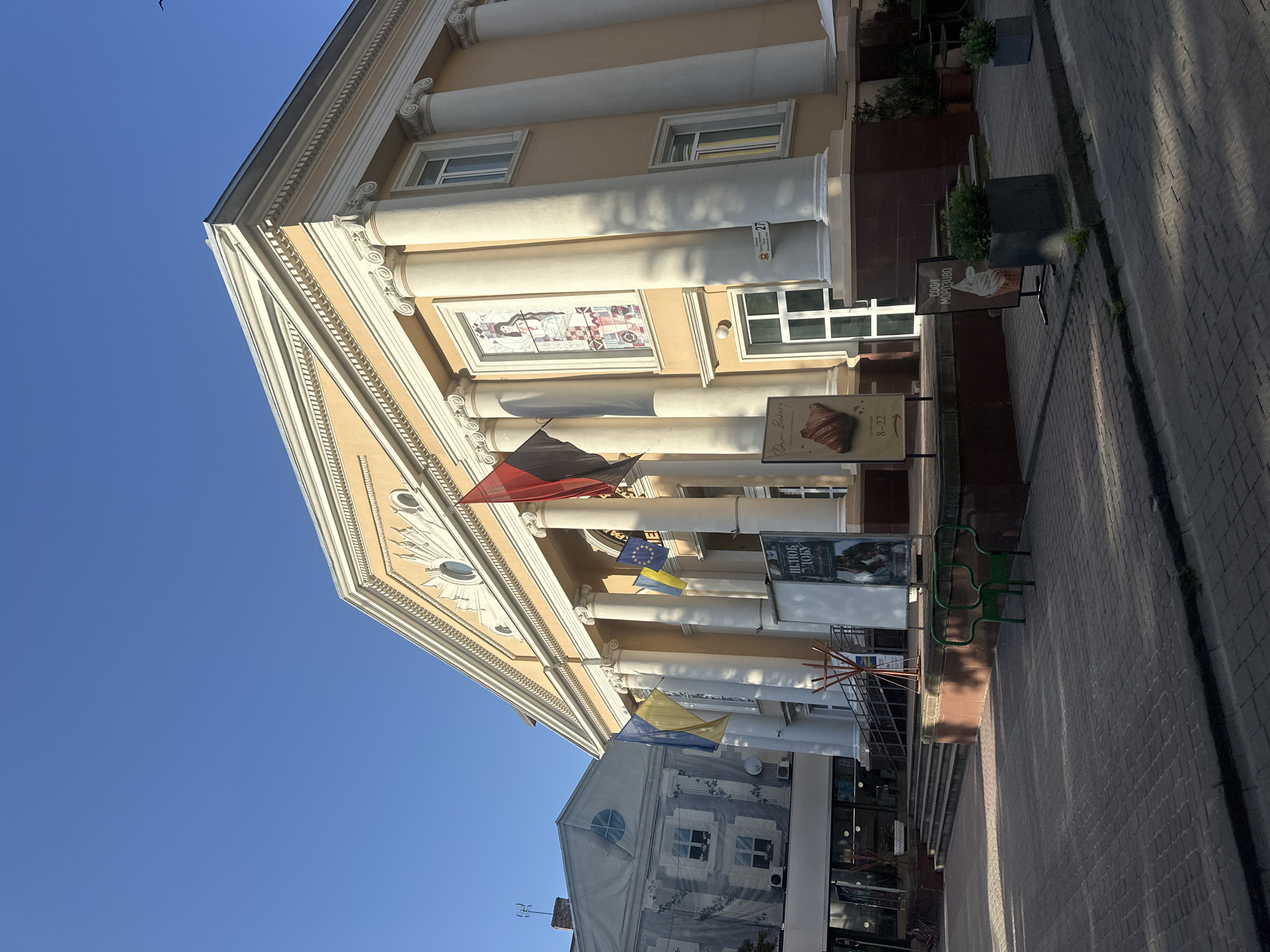 The image depicts the exterior of a building with classical architectural elements, including large columns and a triangular pediment. The building is painted a light beige color and features several windows. Several flags are visible, including Ukrainian flags and what appears to be the flag of the European Union. There are also signs in front of the building, one of which advertises "Dan Bakery" with an image of a croissant and another with an image of ice cream. The scene appears to be outdoors on a sunny day, as indicated by the clear blue sky and the shadows cast on the paved sidewalk. The building is situated on a street with a cobblestone pavement. The overall impression is one of a public or commercial building in an urban setting, likely in Ternopil, Ukraine, given the provided location context. There is a small bird flying in the sky. The image is taken from a low angle, looking upwards at the building.