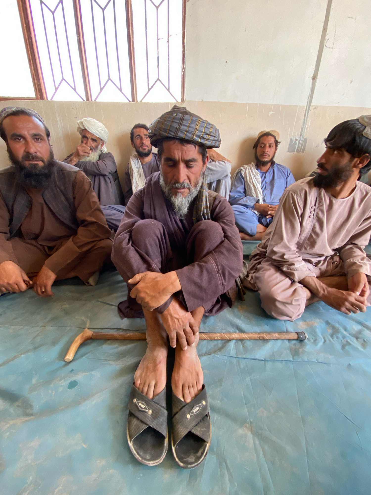 A group of men, appearing to be in their late 20s to 60s, are sitting on a blue tarp in what appears to be an indoor setting, possibly a communal hall or waiting area, in Pasab, Afghanistan. The men are all dressed in traditional clothing, including shalwar kameez and turbans, and some wear vests. One man in the foreground is seated with his legs crossed, wearing black sandals and a checkered turban. He has a beard and is looking down. A wooden walking stick with a curved handle is placed on the tarp in front of him. Other men are seated behind him, some looking towards the camera, others looking away. One man in the background wears a white turban and is gesturing with his hand. The walls behind the men are a neutral color, and there are windows with a lattice design at the top of the frame. The lighting is natural and appears to come from the windows.