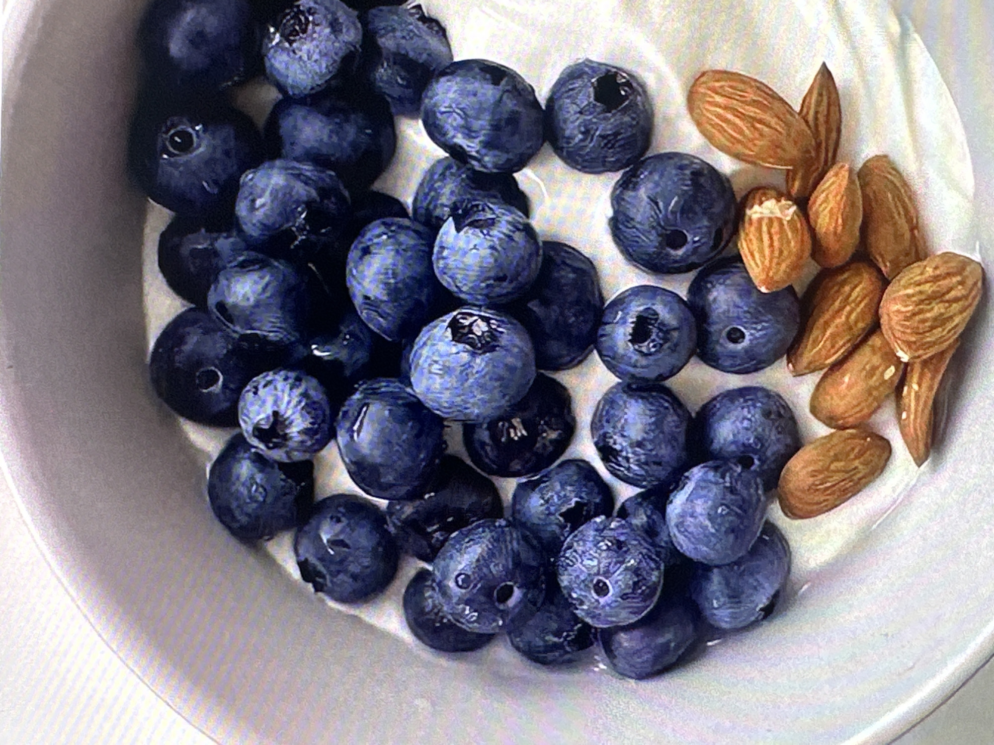 The image shows a white bowl filled with blueberries and almonds. The blueberries are deep blue and clustered together on one side of the bowl. Almonds are arranged on the opposite side. The bowl's contents appear to be mixed with a white substance, possibly yogurt. The location is not discernible from the image.