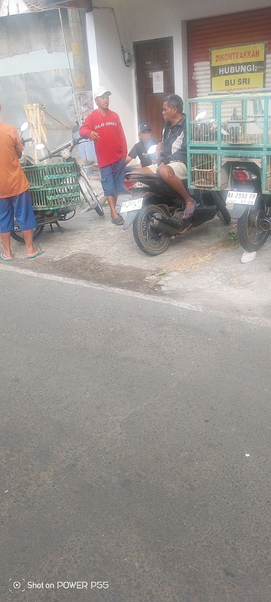 The image shows a street scene with several people and motorcycles. In the foreground, a black motorcycle is parked. To the left of it, another motorcycle is partially visible, with a green cage attached to its back. Three men are standing or sitting near the motorcycles. One man, wearing a red long-sleeved shirt and blue shorts, stands near the green-caged motorcycle. Another man, wearing a black jacket and light-colored shorts, is seated on the black motorcycle. A third man, wearing a black t-shirt and dark pants, is also near the motorcycles. In the background, there is a building with a closed shutter and a sign that reads "DIKONTRAKKAN HUBUNGI: BU SRI". The ground is paved with asphalt, and there is some debris scattered around. The lighting suggests it is daytime.