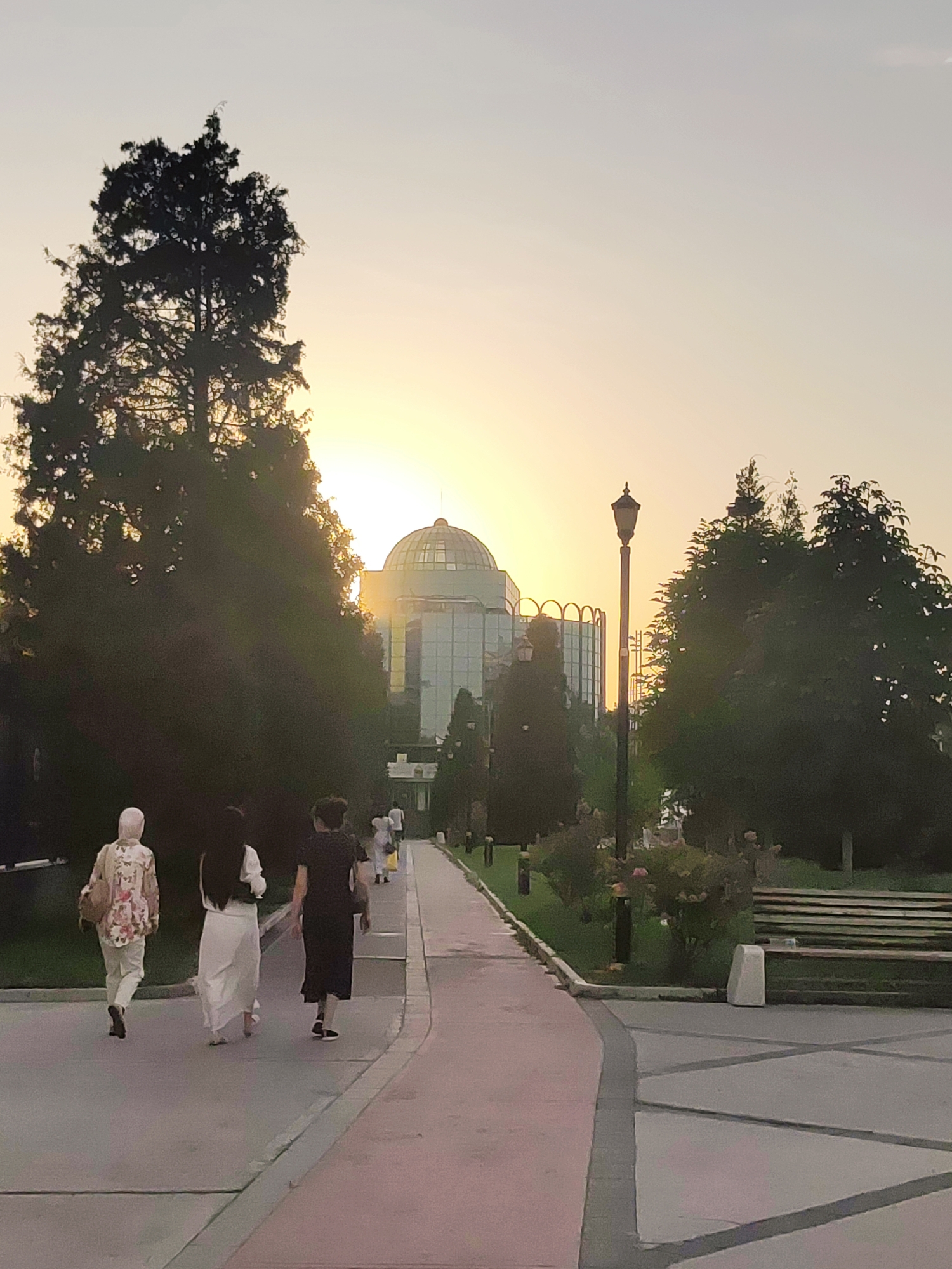 The image depicts a sunset scene at the State Conservatory of Uzbekistan in Tashkent. Three individuals are walking away from the camera on a paved pathway. The person on the left wears a floral patterned shirt and white pants with a headscarf. The middle person is in a long white dress, and the person on the right is in a black dress. A street lamp stands to the right of the pathway. Behind the pathway, trees flank a modern building with a domed roof and glass facade, partially obscured by the setting sun. The sky is a pale orange and yellow gradient. To the far right, a park bench is visible on a grassy area.