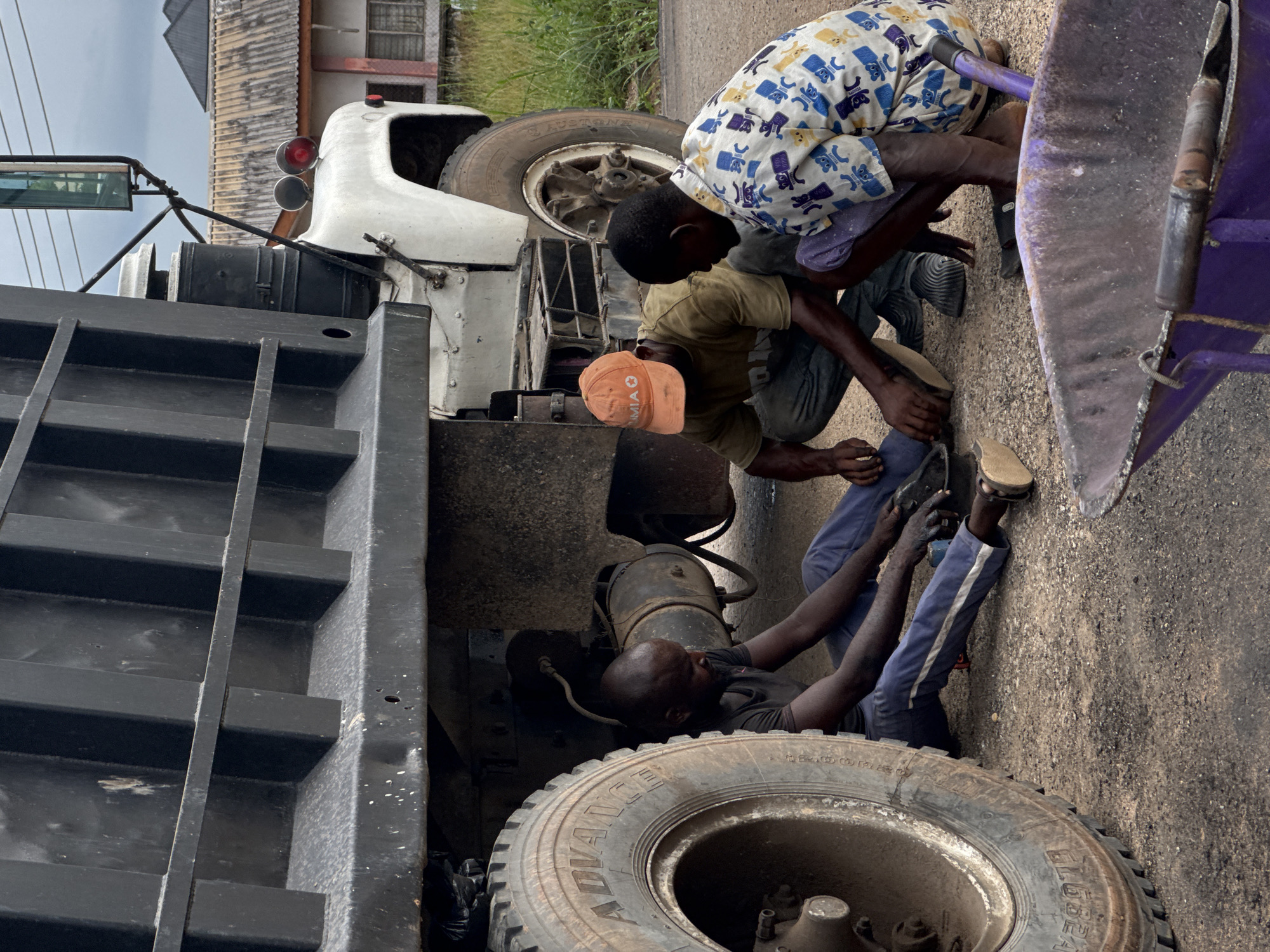 The image captures three men engaged in the repair of a large truck on a paved surface, likely a roadside in Ihiagwa, Nigeria. The scene unfolds during the daytime under an overcast sky.
In the foreground, a large truck tire is prominent, with the words "ADVANCE" and "GLOBAL" visible on its sidewall. One man, dressed in a dark t-shirt and blue pants with white stripes, is lying on his back underneath the truck, actively working on its components.
Two other men are crouching beside the truck. One wears a white and blue patterned shirt, while the other has a light brown t-shirt and an orange baseball cap with "MIA" partially visible. They appear focused on their tasks, with their hands busy near a purple metal object that might be part of the truck's machinery.
The truck itself appears to be a dump truck, with its large black bed partially visible on the left and a white cab section seen towards the top. In the background, simple buildings and some greenery are visible, characteristic of a semi-urban or rural setting. The overall image conveys a dedicated, hands-on repair effort.