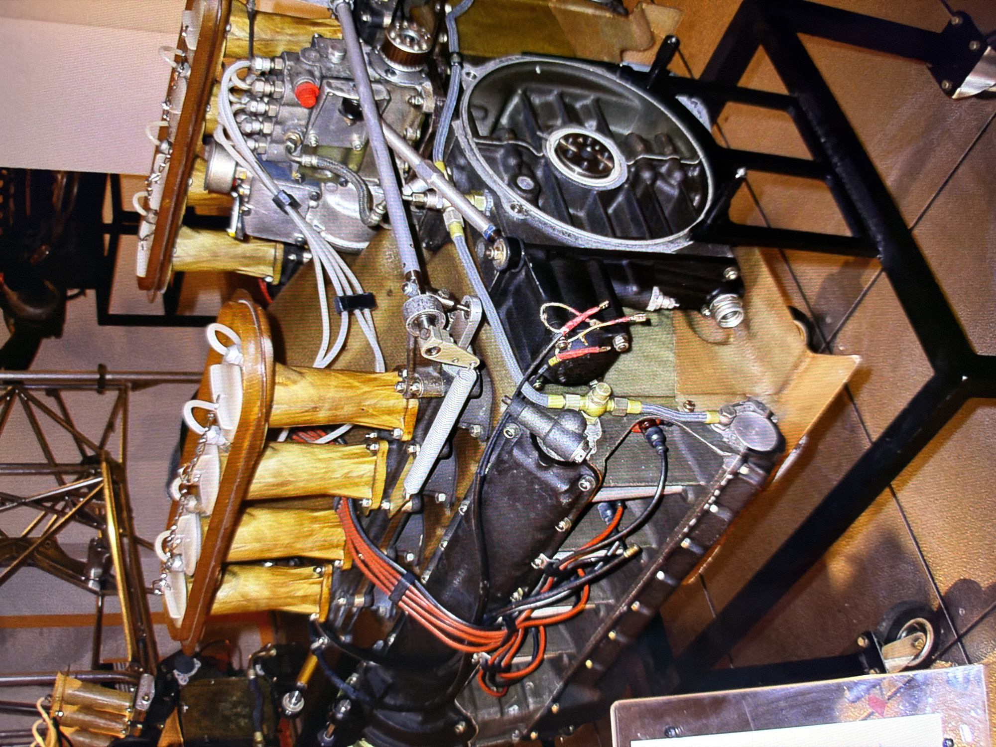 The image depicts a close-up view of a complex engine assembly, likely on display in a museum or exhibition. The engine features multiple intake manifold tubes with wooden casings, connected to what appears to be a fuel injection system. Various metal components, hoses, and wiring are visible, indicating a sophisticated mechanical structure. The engine is mounted on a black metal stand with casters. The background suggests an indoor setting with tiled flooring. The engine itself is the primary subject, occupying the majority of the frame. The scene is static, with no people or actions recorded. The lighting is ambient, highlighting the intricate details of the engine.