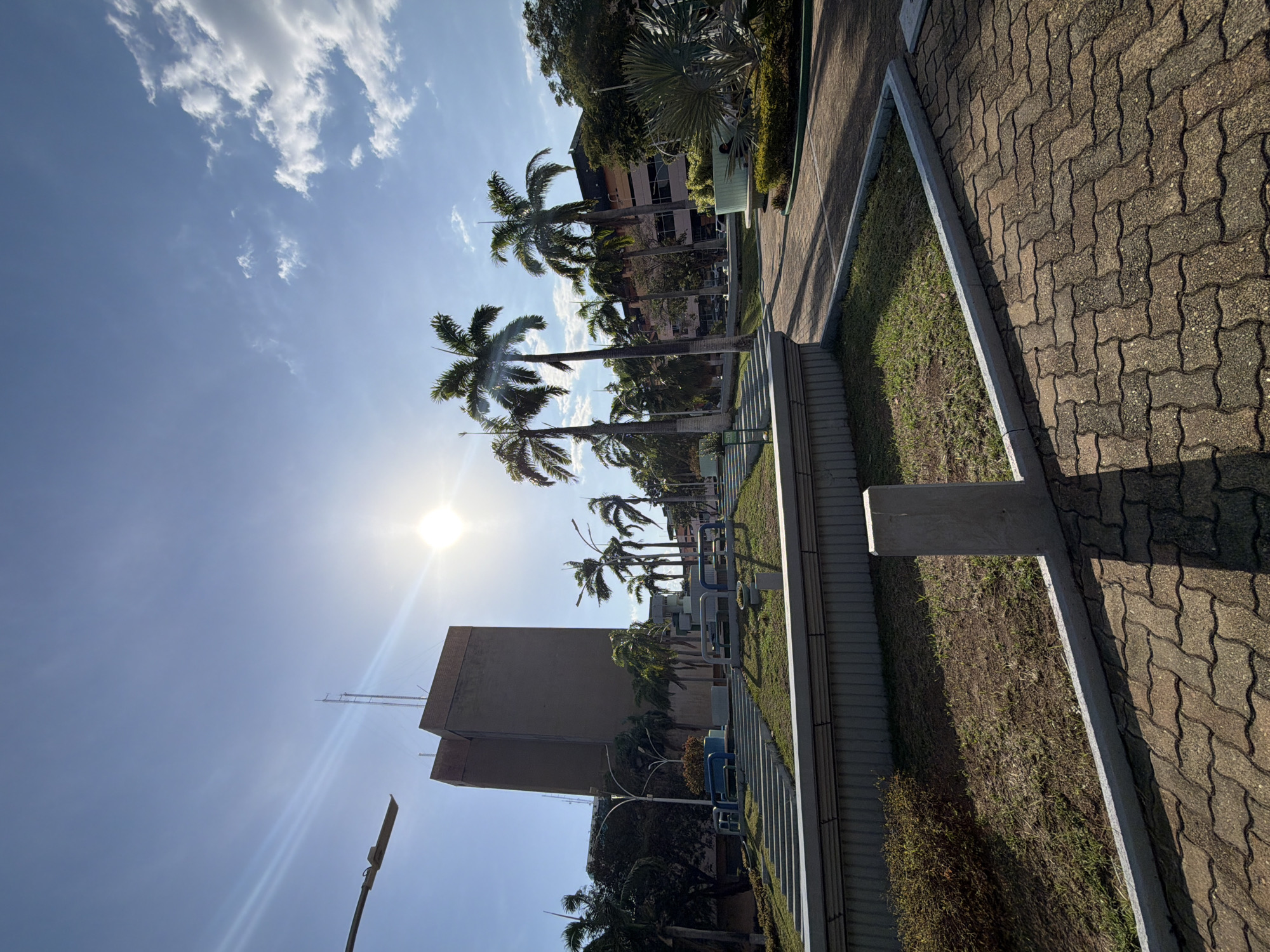 The image shows a street view in Ciudad Guayana, Venezuela, with palm trees lining a paved walkway. The sun is bright in the sky, partially obscured by the palm trees. A building with a square tower is visible in the background, as well as some benches and grass. The pavement consists of light brown interlocking pavers. The sky is blue with scattered clouds.