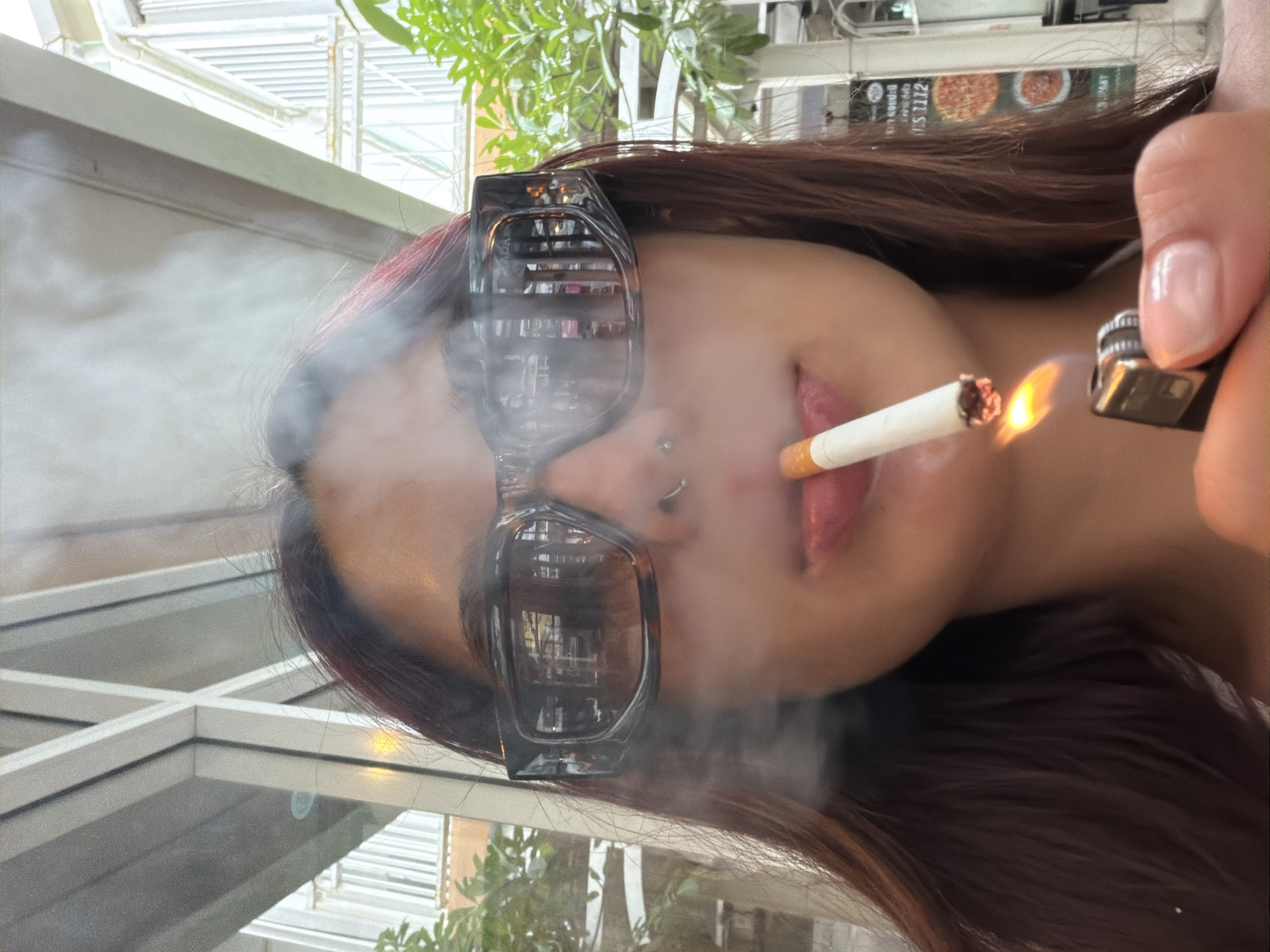The image shows a close-up of a young woman with long, dark hair, wearing oversized, tortoiseshell sunglasses and a nose ring. She is holding a lit cigarette to her lips, with smoke emanating from it. Her right hand is visible, holding a black lighter with a flame igniting the cigarette. The background appears to be an outdoor seating area, possibly a cafe or restaurant, with glimpses of greenery and architectural elements suggesting an urban environment. There is a sign in the background that seems to advertise food, with images of pizzas. The overall lighting suggests it is daytime. The location is identified as Choeng Thale, Thailand.
