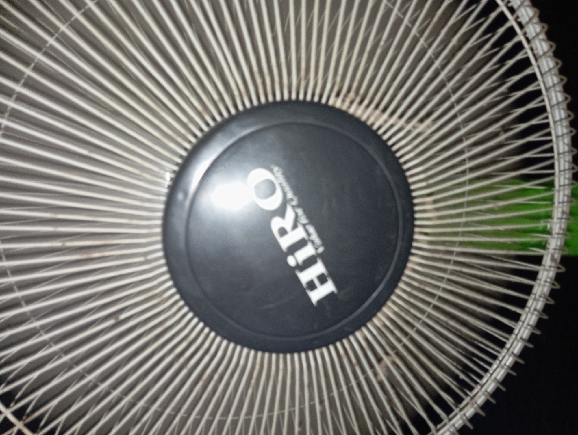 A close-up, angled shot captures the central part of a fan grille, with a black circular emblem prominently displayed. The grille consists of numerous thin, radiating metallic spokes against a dark background. The emblem features white lettering that spells "HiRO" in a stylized font, with "Value for Quality" written below in smaller text. The image is slightly blurry, suggesting a handheld capture or motion. No people or specific locations within Rejosari, Indonesia, are discernible. The overall scene is focused on the fan's design and branding.