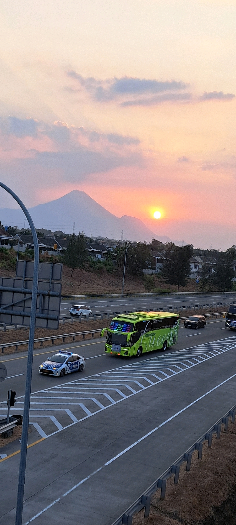 The image depicts a sunset over a highway in Sidowayah, Indonesia. A bright orange sun is partially obscured by mountains in the background. The sky is a gradient of pale yellow and pink, with scattered clouds. In the foreground, a multi-lane highway stretches across the frame. Several vehicles are visible on the highway, including a bright green bus with colorful lights on its front, a white police car with blue and white markings, and other cars. The highway is bordered by a guardrail and dry grass on the right, and by a slope with houses and trees on the left. A large metal pole with signage is present on the left side of the image. The scene appears to be a snapshot of daily life on a busy road during a beautiful sunset.