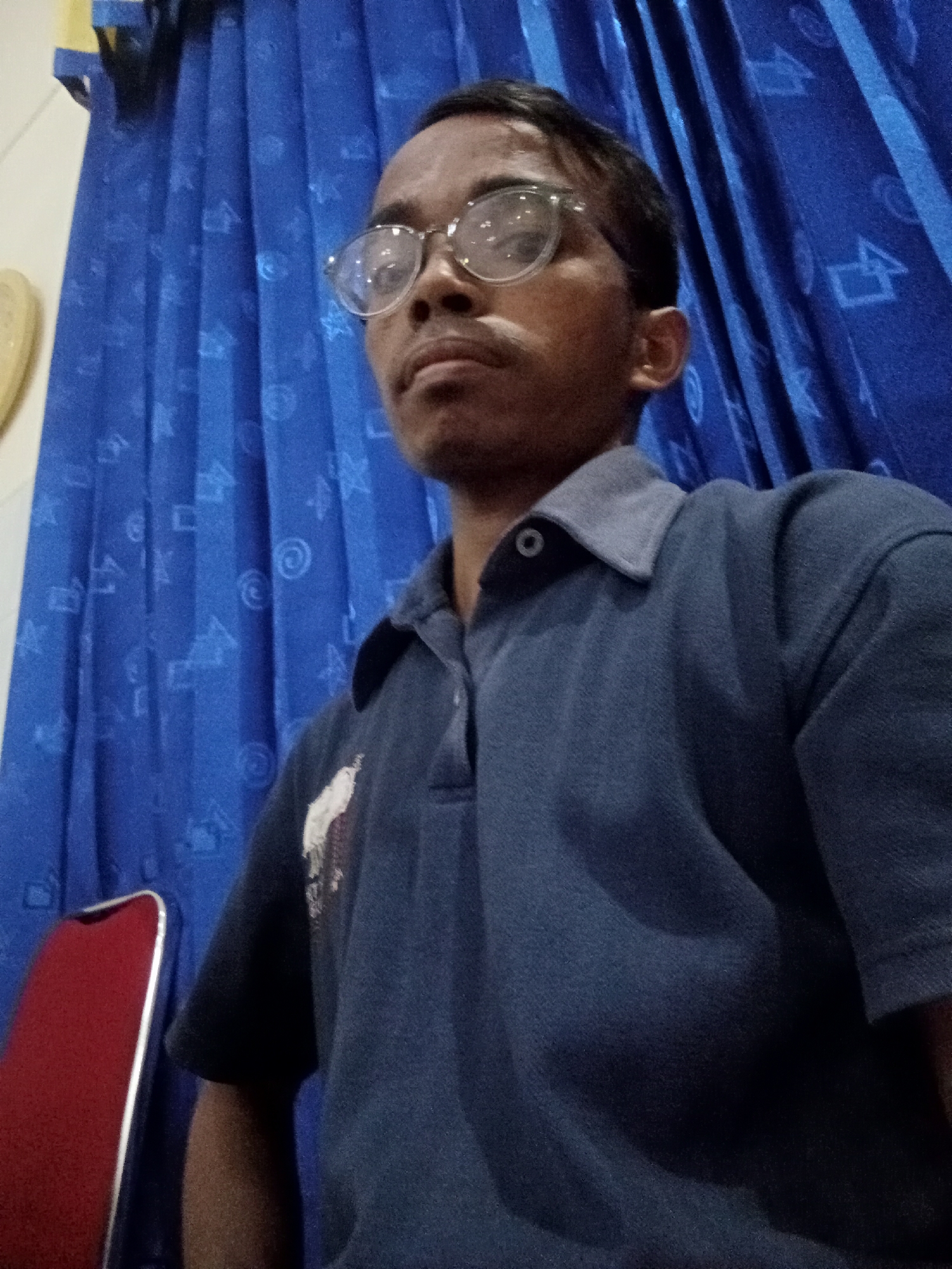 A male is seen in a selfie, wearing a blue polo shirt and clear-rimmed glasses. He is positioned in front of a blue curtain with a pattern of white shapes. To the left of the frame, a portion of a red chair with a silver trim is visible. The lighting appears to be artificial. The image was taken indoors. The user's location is indicated as Surabaya, Indonesia.