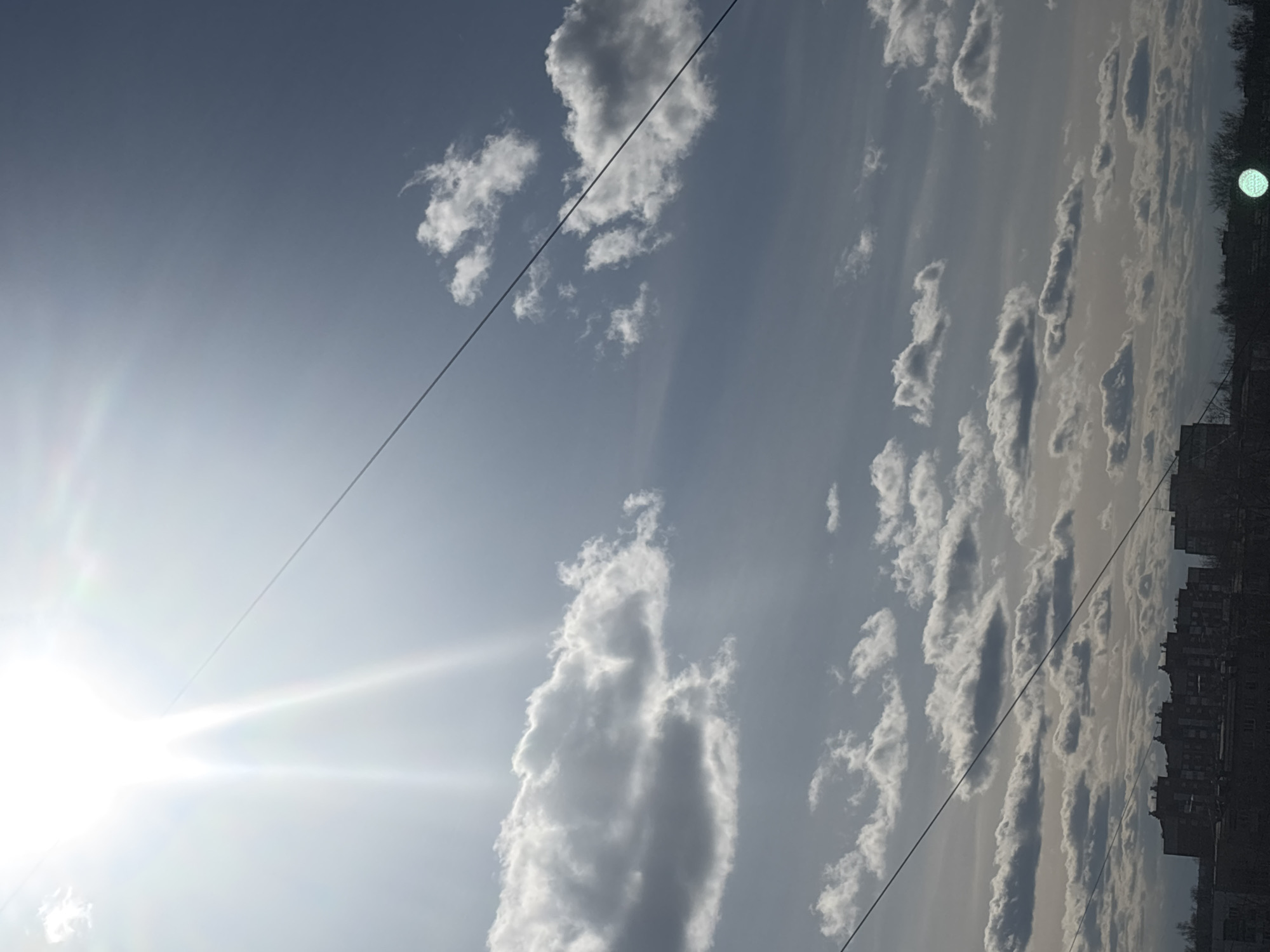 The image depicts a daytime sky in Chaikovsky, Russia, characterized by scattered cumulus clouds. A bright sun is positioned on the left side of the frame, its glare obscuring details in that area. Two dark wires bisect the sky diagonally. Along the right edge of the image, indistinct buildings and trees form the city's skyline. A prominent bright green spot is at the top right corner.