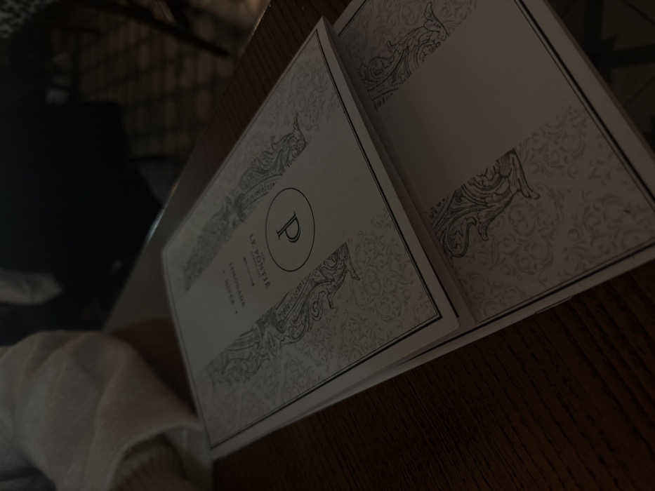 An open menu sits on a wooden table. The cover features a decorative design with a large letter "P" encircled. Below the "P" is text, including the words "LE PONTIE", "LIMONADE", and "RIVIERA". The open pages are mostly blank, framed by similar decorative elements as the cover. A white fabric is visible at the bottom left corner of the frame.