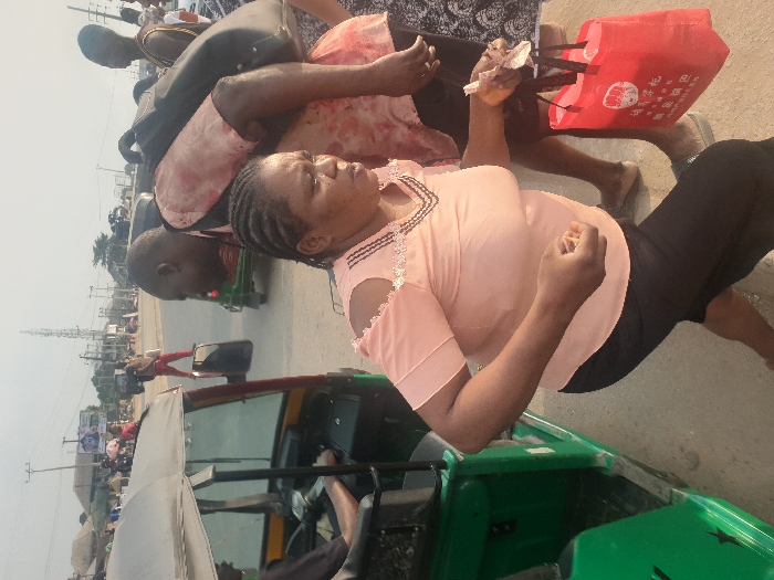 A woman in a pink top and black pants is standing on a street near a green motorized tricycle. She holds tissues in one hand. People pass behind her, with a red shopping bag visible. A man with a black backpack is partially visible in the background. The tricycle is parked on the side of a paved road, and a driver is seen sitting inside. Power poles and buildings are visible in the distance under a bright sky.