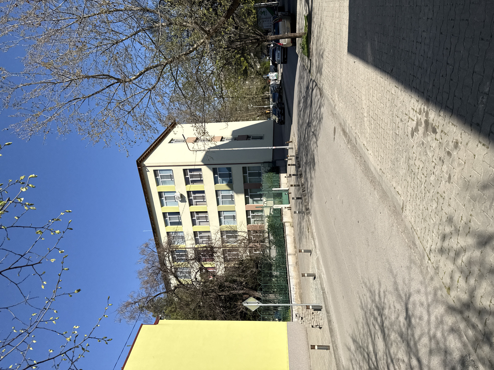 The image shows a street scene in Stara Zagora, Bulgaria, under a clear blue sky. The perspective is rotated counter-clockwise. A five-story building with a beige facade and brown roof is prominent on the left. Each floor features multiple windows with white frames. The building is partially obscured by tree branches, some with fresh spring buds.
Adjacent to the tall building, a shorter, one-story yellow building is visible. A green metal fence runs along the perimeter of the buildings. The street is paved with gray asphalt and cobblestones. Shadows from trees are cast on the cobblestone sidewalk. A row of parked cars is on the right side of the street in the distance. A street sign with a diamond shape is visible near the shorter building.
