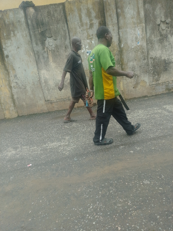 Two males are depicted walking leftward on a paved road surface adjacent to a weathered concrete wall in Agani, Nigeria.
The individual in the foreground (right) is a male wearing a short-sleeved green and yellow t-shirt with partial text, black athletic pants with a white vertical stripe, and dark Crocs-style sandals. A dark strap or belt hangs from his right hip.
The individual slightly behind and to the left is a slender male wearing a dark short-sleeved shirt, dark shorts, and dark flip-flops. He is carrying a small, multi-colored object (red, blue, metallic) with a chain in his right hand. Both individuals appear to be moving in the same direction.
The ground is a dark grey asphalt or concrete surface, unevenly covered with light brown dust and small aggregates. A small piece of red and white debris is visible on the road surface in the lower-left quadrant. The background consists of a tall, light-colored concrete wall, segmented vertically, with a circular embossed design in the upper-left portion and visible remnants of faded posters or paper adhered to its surface. The wall exhibits signs of weathering and discoloration. The scene is illuminated by ambient daylight.