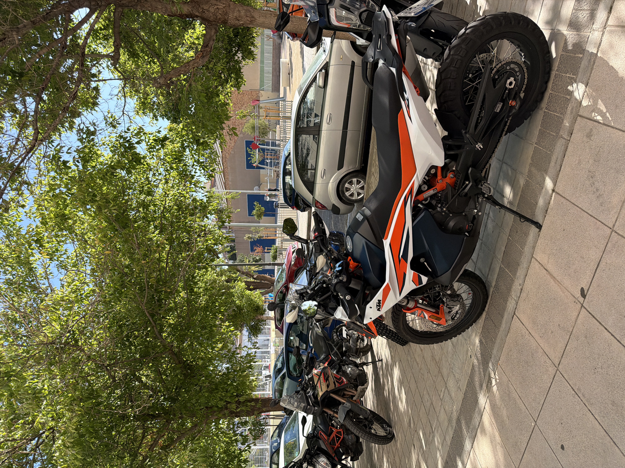 In an outdoor urban setting, several motorcycles and cars are parked on a paved area under the shade of mature trees. The foreground features a white and orange KTM motorcycle, angled to the right, with its kickstand down. To its left and behind, other motorcycles are visible, including one with orange and black accents. Several cars are parked in the background, including a silver hatchback and darker sedans. The environment is sunny, with dappled light filtering through the green leaves of the trees. In the far background, buildings and playground equipment can be seen, suggesting a park or public area within the city.