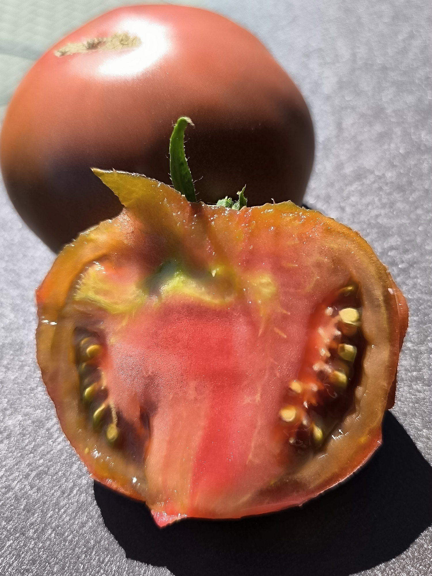 A close-up, overhead shot shows a dark red, whole tomato behind a tomato cut in half. The cut tomato reveals its red flesh, yellow seeds, and green stem. The tomatoes rest on a textured, gray surface, possibly a table, with a shadow cast from the whole tomato. The lighting suggests an outdoor setting with natural sunlight. There are no people present in the image, and no specific location in Dercekliai, Lithuania can be identified from the visual information provided.