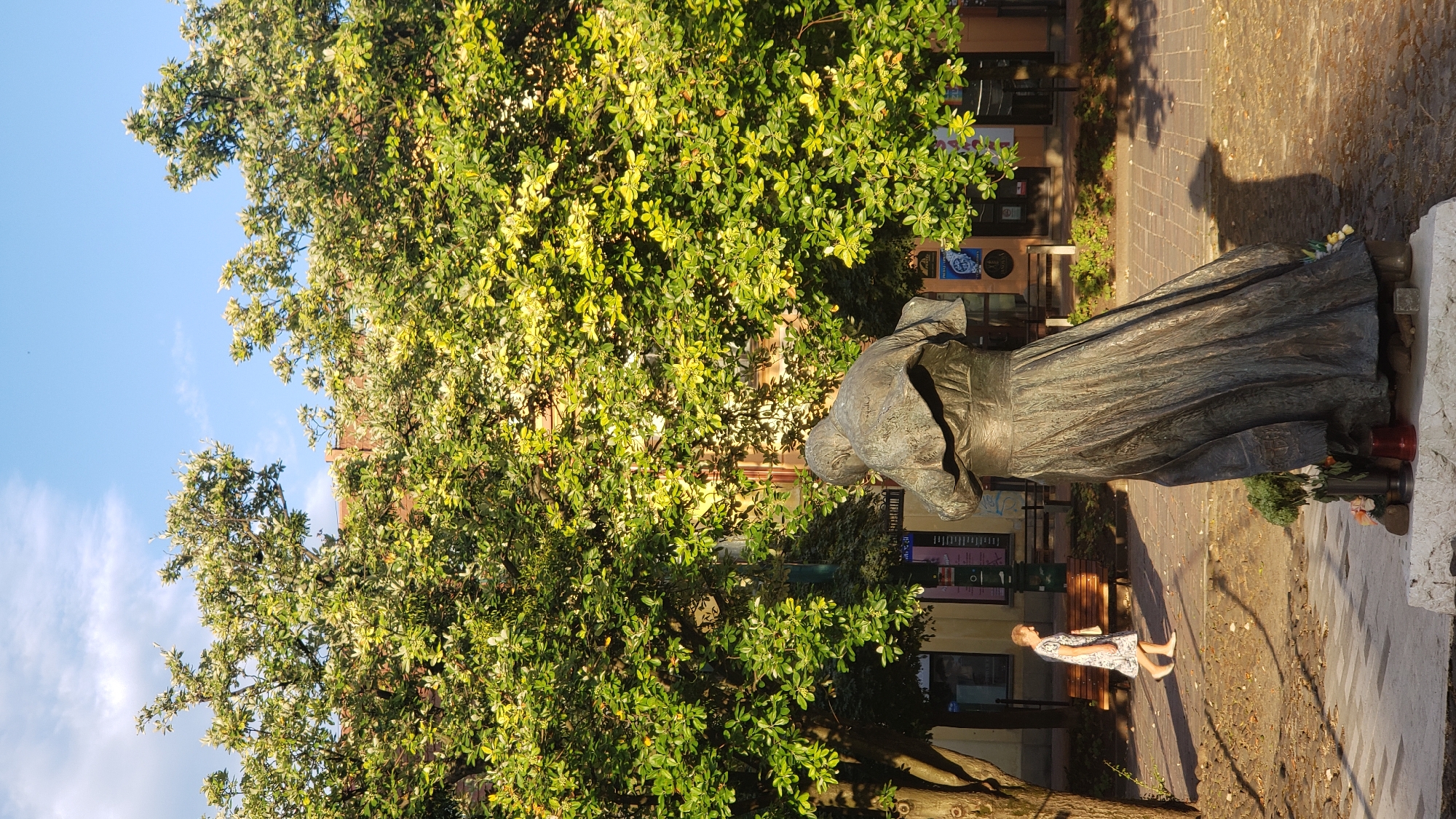 The image shows a bronze statue of a cloaked figure in a public square. The statue is situated in front of a row of buildings and is surrounded by large trees with lush green leaves. A person, a woman wearing a light-colored dress, is walking on a paved walkway in the background, to the left of the statue. The ground is paved with cobblestones and some decorative stones. The sky above is blue with a few white clouds. The lighting suggests it is late afternoon or early morning, with long shadows cast by the trees and the statue. The overall scene depicts a tranquil urban setting. This location is likely the main square of Prešov, Slovakia, given the context of public art and urban architecture often found in such areas.