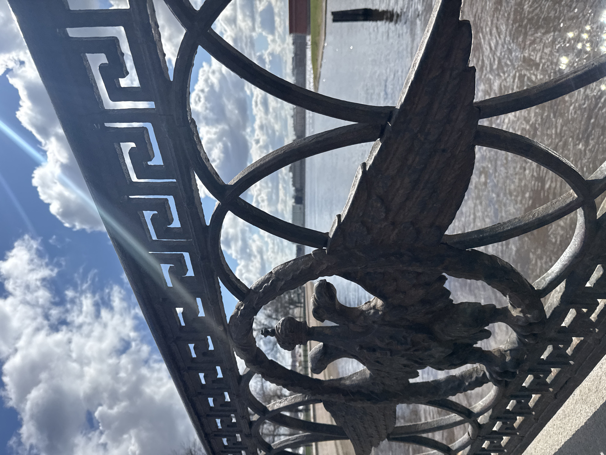 The image is a vertical-oriented close-up shot of an ornate dark metal railing, likely part of an embankment or bridge. The railing features two distinct decorative elements: along its upper edge, there is a repeating geometric pattern resembling a Greek key or meander; at its center, a prominent intricate design depicts a two-headed eagle. This eagle, characteristic of Russian heraldry and specifically the Romanov imperial eagle, is shown with spread wings, clutching a scepter and an orb, and adorned with crowns.
Through the gaps in the metalwork, a wide body of water—possibly a river or canal—is visible, reflecting the bright, cloudy sky. Distant land or structures can be faintly discerned across the water. The sky is a clear blue with scattered white, cumulus clouds, indicating daytime conditions. A distinct lens flare emanating from the sun is visible in the upper left of the frame, highlighting the bright, sunny weather. The metal of the railing appears textured and aged.
Given the provided location context of Saint Petersburg, Russia, the presence of the two-headed eagle and the classical architectural decorative style strongly suggest this is a detail from a historical structure such as a bridge, a palace fence, or a riverside embankment in the city. There is no specific activity or event depicted, and no visible text.