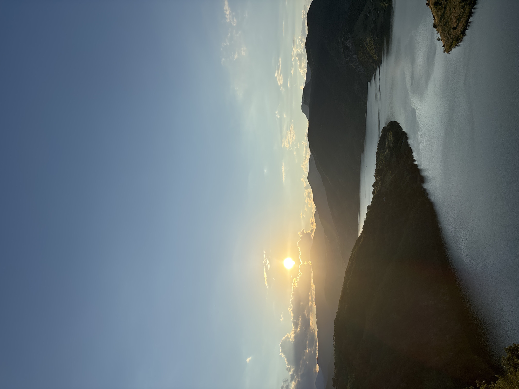 This is a landscape photograph taken at sunset in the vicinity of Borovitsa, Bulgaria. The image features a body of water, likely a reservoir, winding through a valley surrounded by rolling, tree-covered hills and mountains. The setting sun is visible in the upper left quadrant of the image, partially obscured by clouds, casting a warm glow on the sky and illuminating some of the clouds with a golden hue. The light creates a strong contrast between the silhouetted, dark shapes of the landscape and the illuminated sky. The water reflects the sky and the surrounding scenery, appearing dark and serene. A small, grassy promontory juts into the water on the right side of the frame. The overall scene evokes a sense of tranquility and natural beauty during dusk.