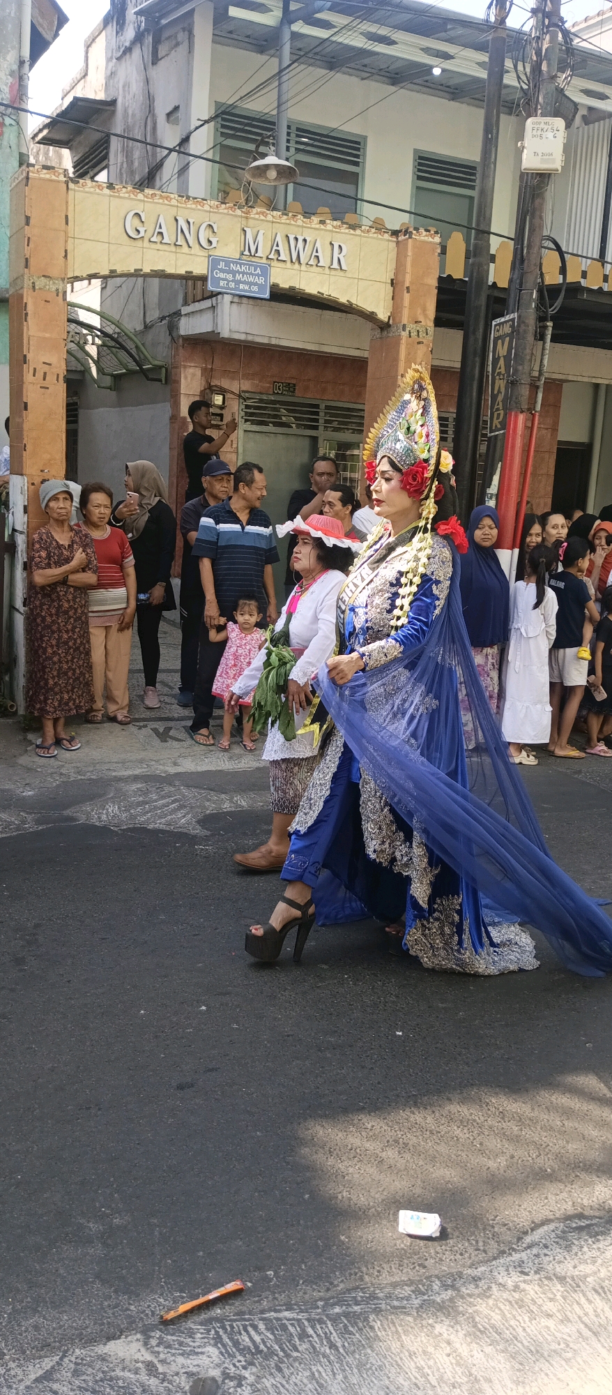 In front of an archway with the text "GANG MAWAR" and a street sign reading "JL. NAKULA GANG. MAWAR RT. 01 - RW. 05", a procession is taking place on a street in Malang, Indonesia. A woman adorned in a traditional blue and silver costume, complete with an elaborate headdress and a long, flowing blue veil, walks towards the right of the frame. She is wearing high-heeled platform shoes and has flowers in her hair and around her neck. She is accompanied by other individuals, including a child in a pink dress, a woman in a red hat, and several onlookers dressed in casual attire. The environment consists of buildings with visible electrical wires and poles, indicating an urban setting. The ground is paved asphalt with some painted lines and debris. The lighting suggests it is daytime.