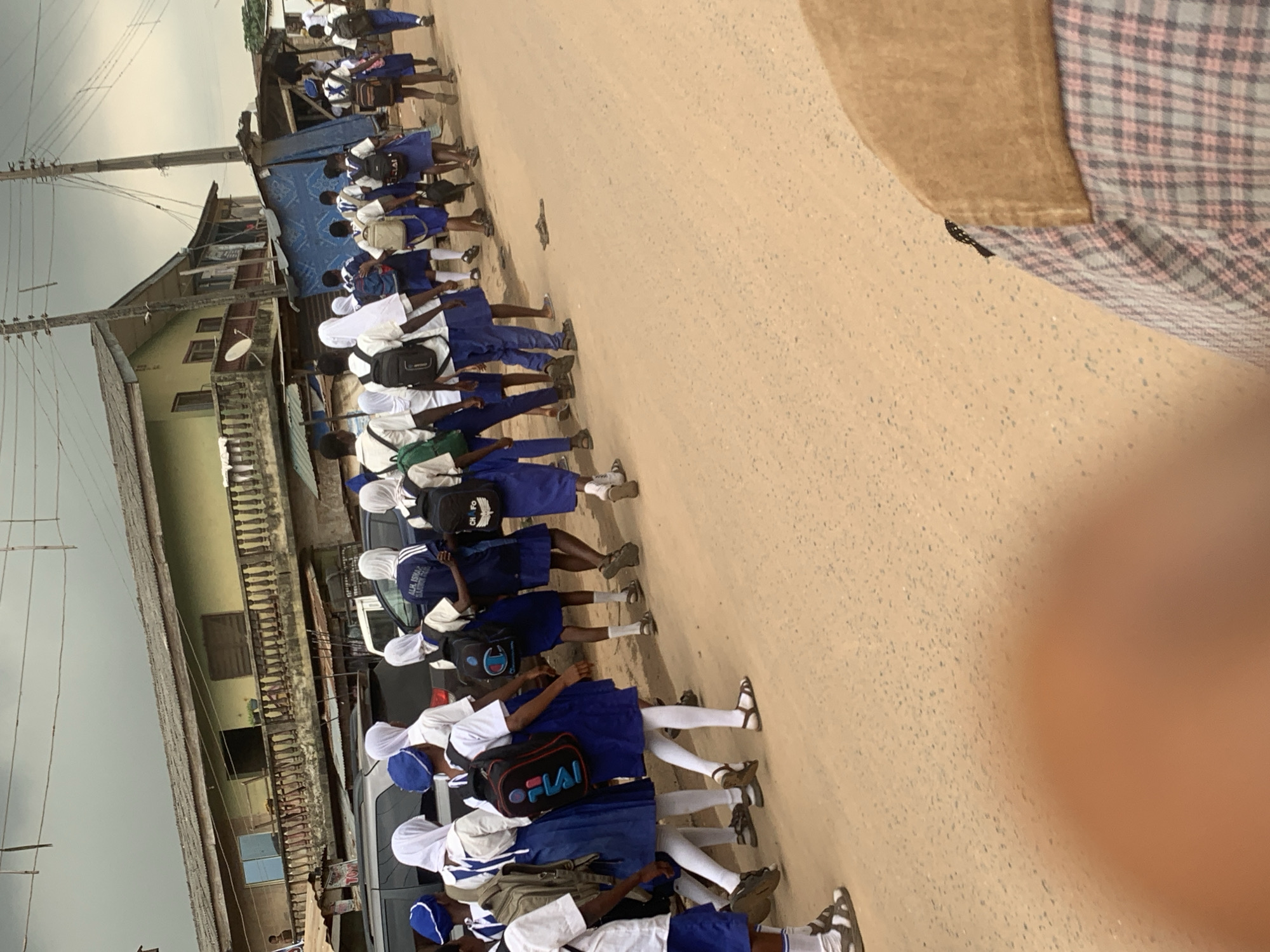 In Shagamu, Nigeria, a group of schoolchildren in blue and white uniforms are walking along a dusty road. They are wearing white headscarves and carrying backpacks. The children are walking in a line, with some children in the front and others following behind. In the background, there are buildings and a street. The sky is cloudy. The image appears to have been taken from a low angle, possibly from a vehicle or from someone walking in the same direction as the children.
