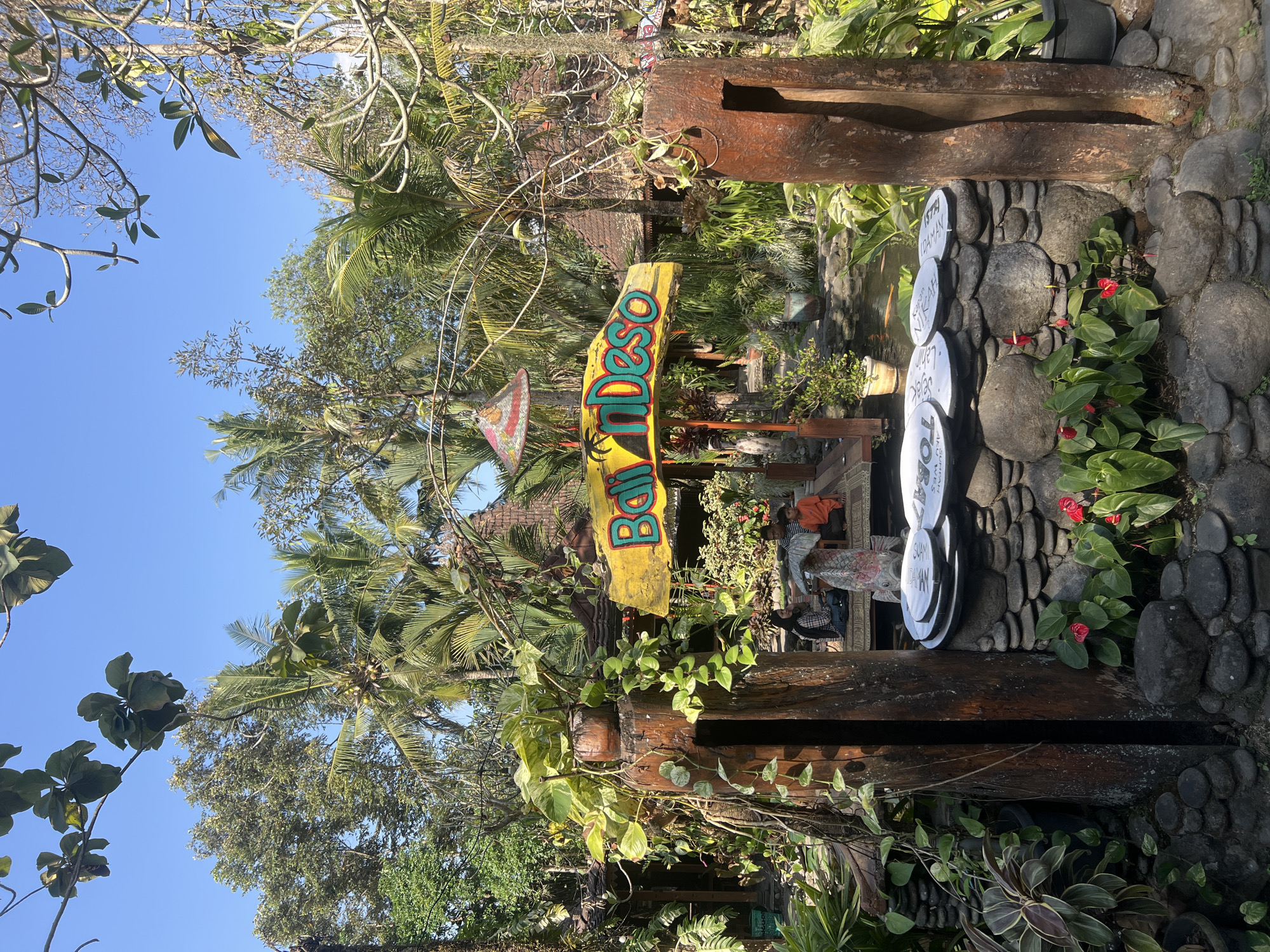 The image depicts an outdoor scene in Tlogoadi, Indonesia, likely a cafe or restaurant. In the foreground, a weathered wooden structure stands on the left, with lush green plants cascading down its sides. To the right of the wooden structure, a stone wall is adorned with several white plates. A bright yellow sign with colorful lettering spelling out "Bali n'Deso" is prominently displayed above a small pond. Behind the sign, several people are visible sitting on a raised platform, appearing to be customers. The background is filled with dense tropical vegetation, including palm trees and other lush greenery, under a clear blue sky. The overall atmosphere is relaxed and tropical, suggesting a leisure setting.