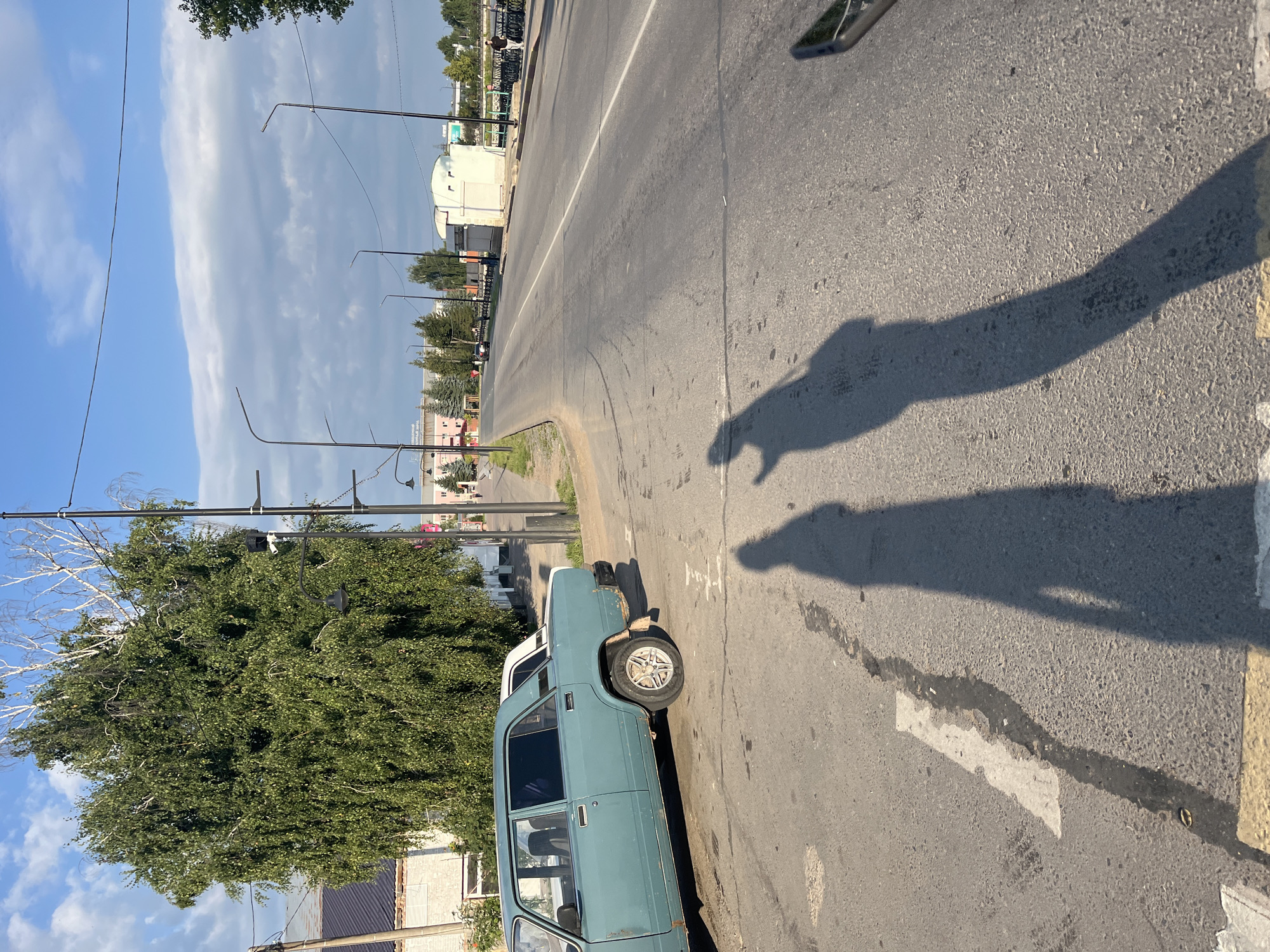 The image depicts a street scene in Novye Turbasly, Russia. A light blue, older model sedan is parked on the left side of the frame, angled towards the street. The car shows signs of rust, particularly on the rear fender. In the foreground, the shadows of two people are cast on the asphalt, with one shadow appearing to be raising an arm. The street itself is paved with asphalt and has clear lane markings. A white line with some dark staining runs diagonally across the road in the foreground. The background reveals a tree-lined street with a sidewalk and a few buildings. Streetlights are visible along the road. The sky above is blue with scattered clouds.