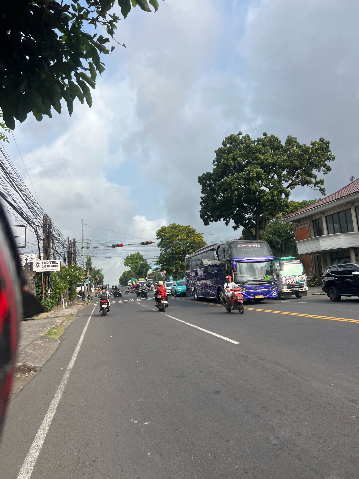 A street scene in Denpasar, Indonesia, depicts a moderate traffic flow on a cloudy day. In the foreground, the edge of a red motorcycle helmet is visible, indicating the perspective is from a rider. The road is paved with asphalt and marked with white and yellow lines. Several motorcycles, a bus, and a truck are moving on the road. On the left side of the road, there's a sidewalk and a building with a sign that reads "HOTEL GATSU INDAH." Utility poles with numerous wires are also present. Further down the road, trees and other buildings are visible. A traffic light is illuminated red. The sky is overcast with large clouds.