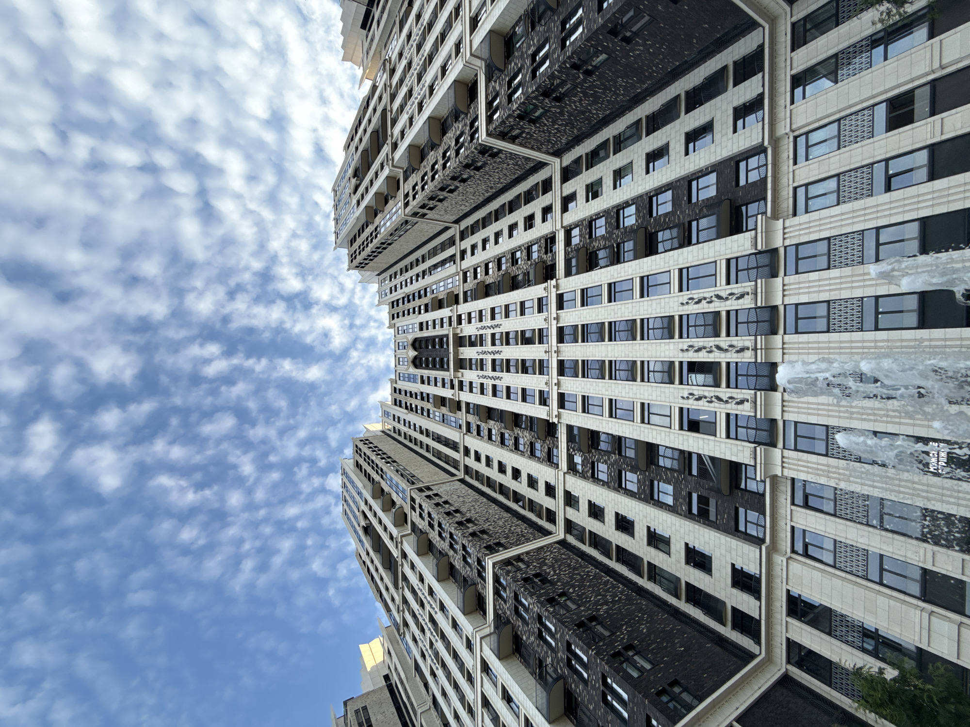 The image shows a low-angle view of a modern residential skyscraper in Moscow, Russia. The building features a facade with alternating light-colored stone and dark brickwork. Numerous windows and balconies are visible, creating a repetitive pattern. In the foreground, a fountain sprays water upwards, partially obscuring the lower part of the building. The sky above is blue with scattered white clouds.
