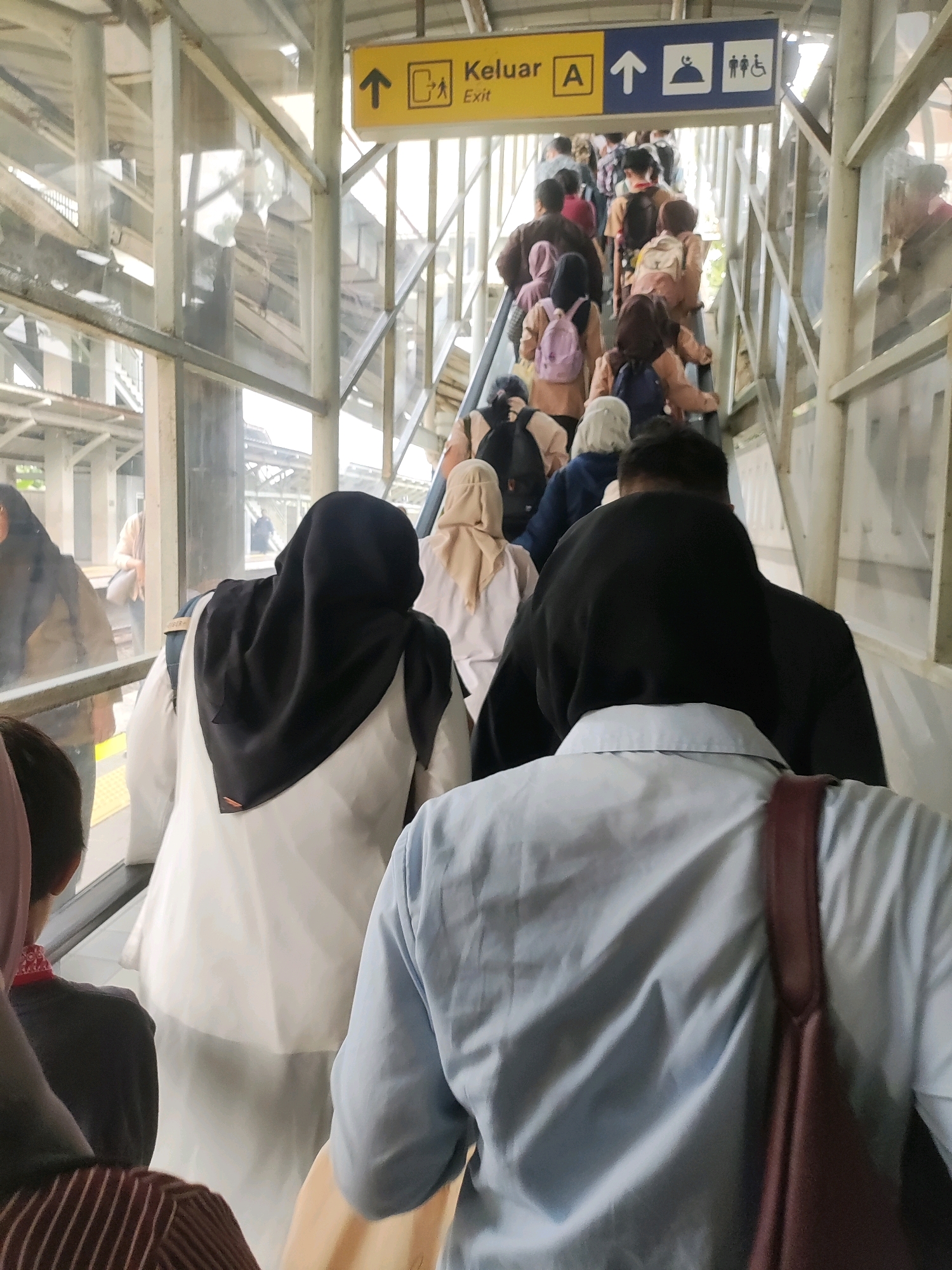 A group of people are ascending an outdoor escalator at a train station in Telagamurni, Indonesia. The escalator is enclosed by a metal structure with glass panels. Signage above indicates "Keluar A" (Exit A) with an arrow pointing upwards and icons for a person, a mosque, and a wheelchair. Most of the people are wearing casual clothing, and many have backpacks. The foreground shows the backs of several individuals, including one wearing a black headscarf and white clothing, and another in a light blue collared shirt. Sunlight filters through the structure, illuminating the scene.
