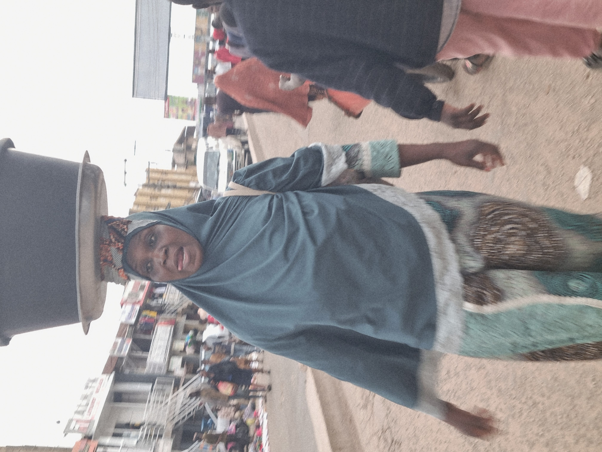 The image depicts a street scene in Jos, Nigeria, with a focus on a woman carrying a large, dark pot on her head. She is wearing a teal hijab and a patterned dress in shades of blue, green, and white. Her mouth is open, suggesting she might be speaking or reacting to something.
The scene appears to be a busy market or street with other people visible in the background. Some are dressed in casual clothing, including a person in pink pants and another in a dark jacket. The background shows buildings with multiple stories and what appear to be shops or stalls, indicating an urban or semi-urban environment. The ground is paved with a sandy or dusty surface.
The lighting suggests it could be daytime, possibly late morning or afternoon, with overcast or diffused light. There are no clear indications of specific weather conditions, but the general atmosphere seems to be that of an active day in a community. The overall impression is of everyday life and commerce taking place on a street in Nigeria. The image is slightly blurry, which adds to a sense of movement and immediacy.